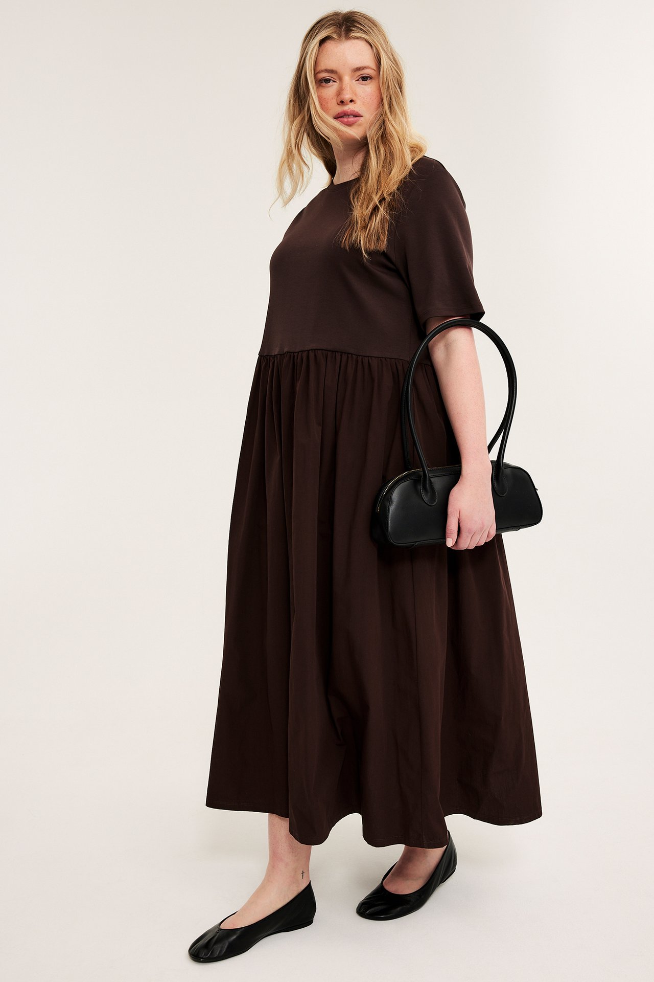 Framifrån, kvinna i mörkbrun rundhalsad klänning med jerseytop, poplinkjol, håller en svart handväska och bär svarta ballerinaskor.
