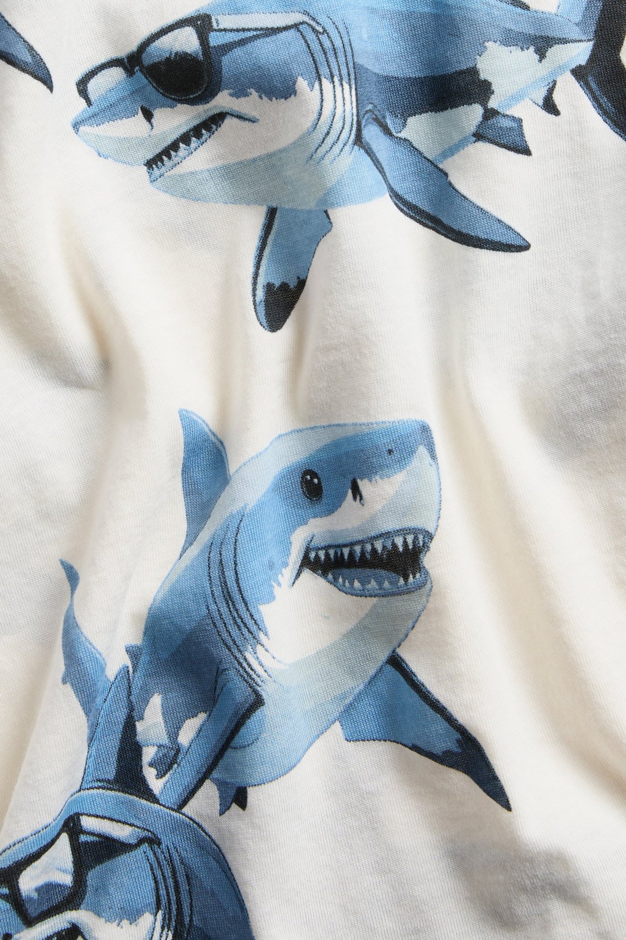 Zbliżenie: dziecięcy T-shirt z białej bawełny z niebieskim nadrukiem rekinów; niektóre rekiny noszą okulary.