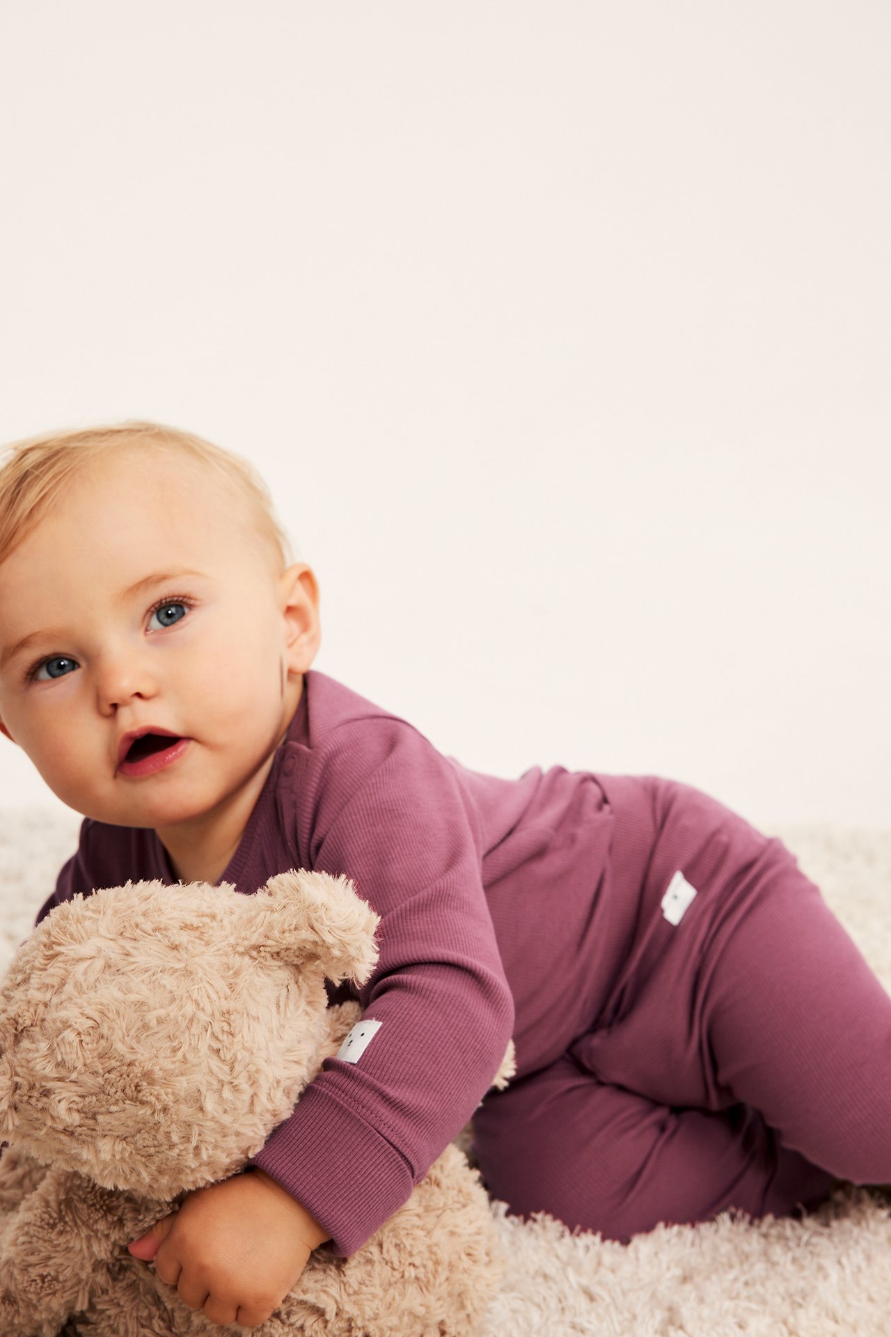 Forfra, baby i plommefarget ribbestrikket langermet body med hvite lapper, holder teddybjørn.