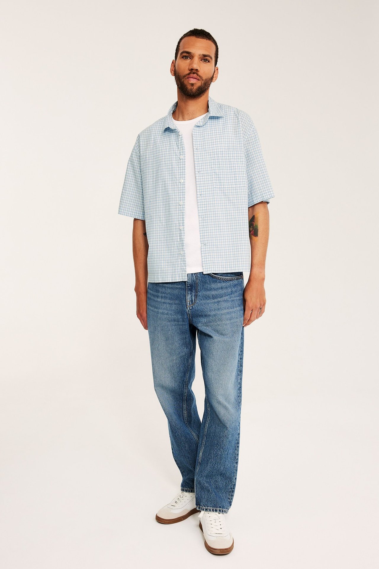 Frontvy: man i öppen ljusblå rutig bomullsskjorta över vit t-shirt, blå jeans och vita sneakers.