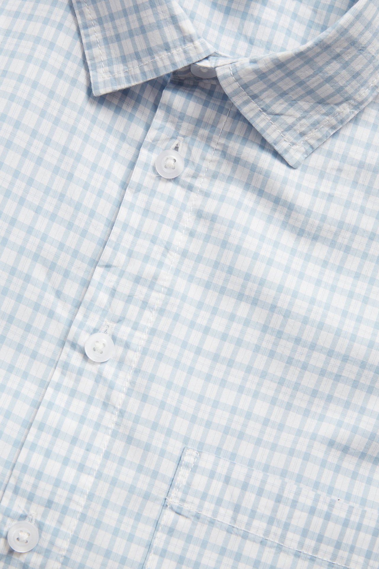 Närbild av en blå- och vitrutig kortärmad herrskjorta i bomull med krage, knappar och bröstficka.