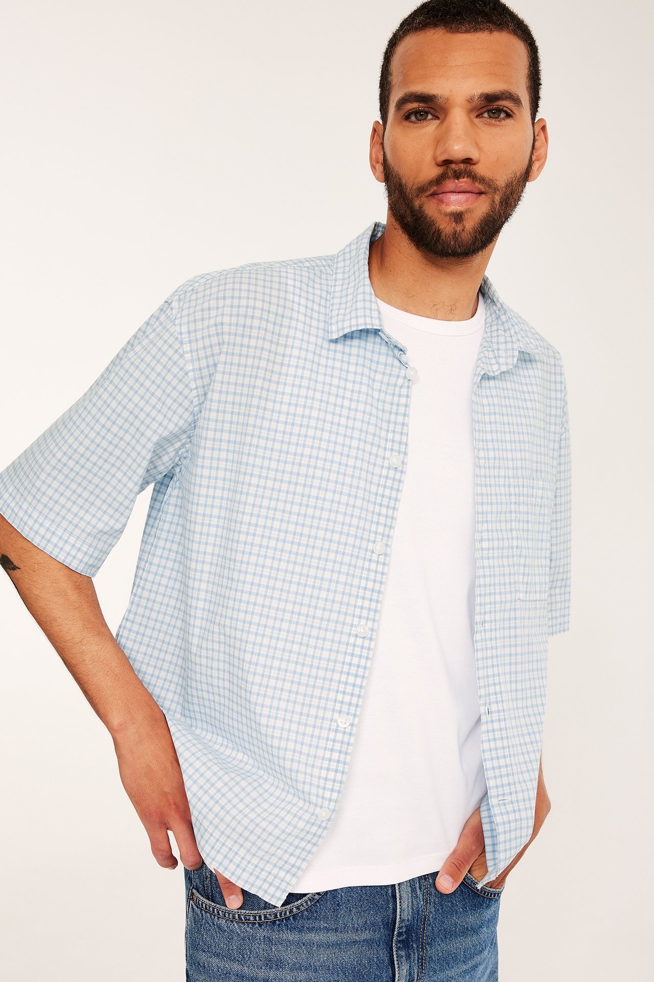 Man i ljusblårutig kortärmad poplinskjorta över vit t-shirt och blå jeans, framifrån.