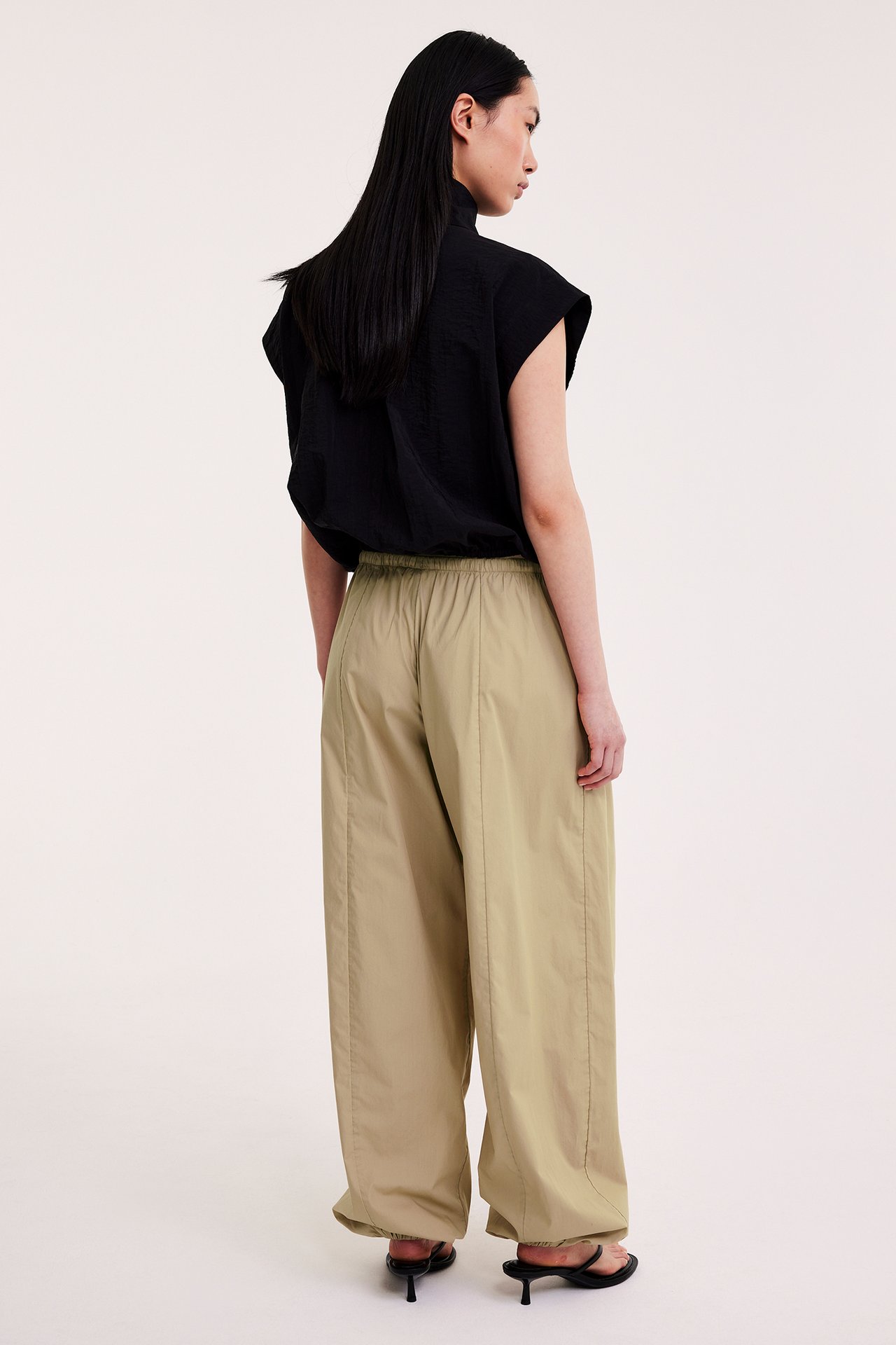 Naisen selkä, beiget puuvillaiset ballongihousut resårilla, musta toppi ja mustat korkokengät sandaalit.