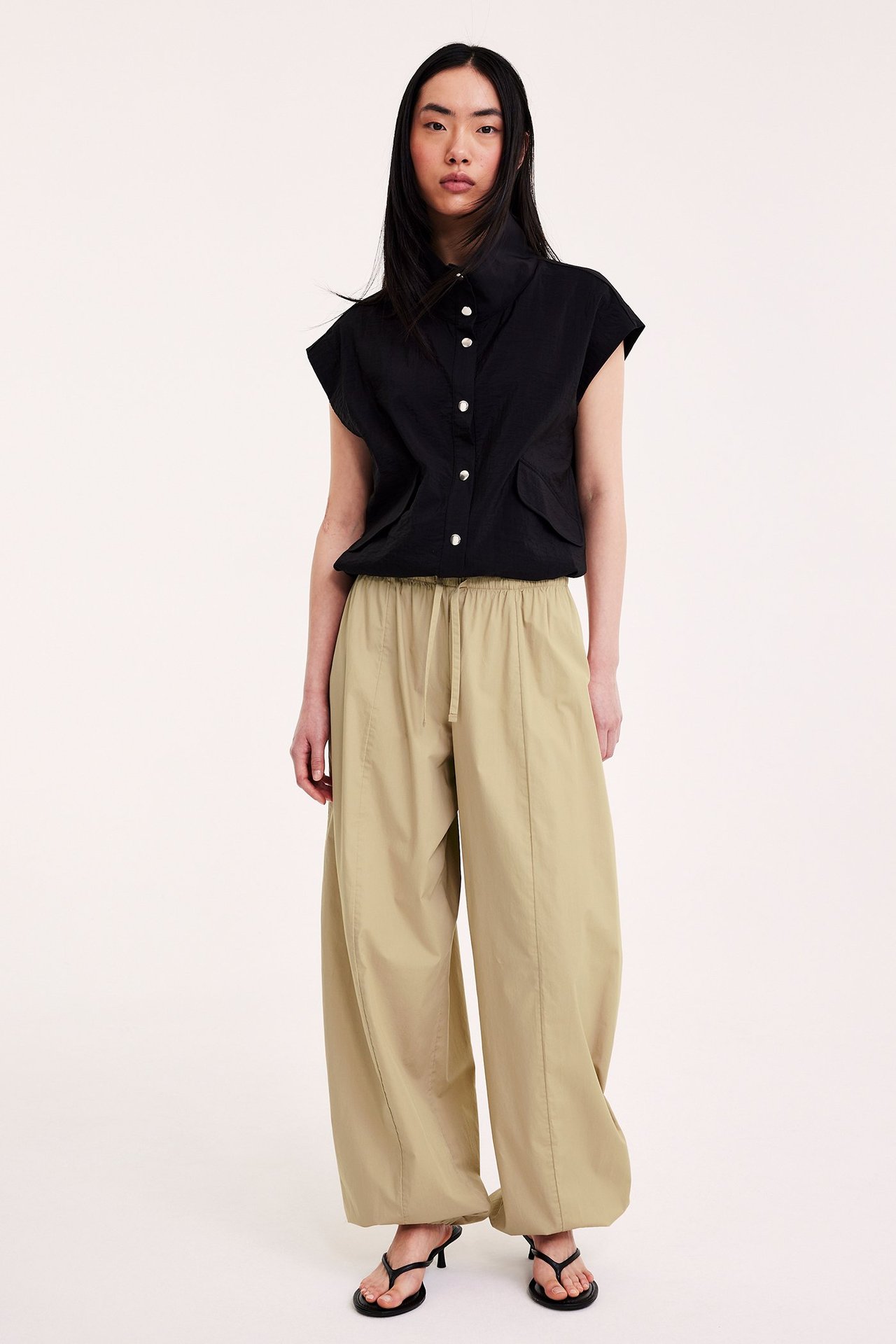 Framifrån en kvinna i beiga bomullsballongbyxor, en svart ärmlös kragskjorta och svarta klackade sandaler.