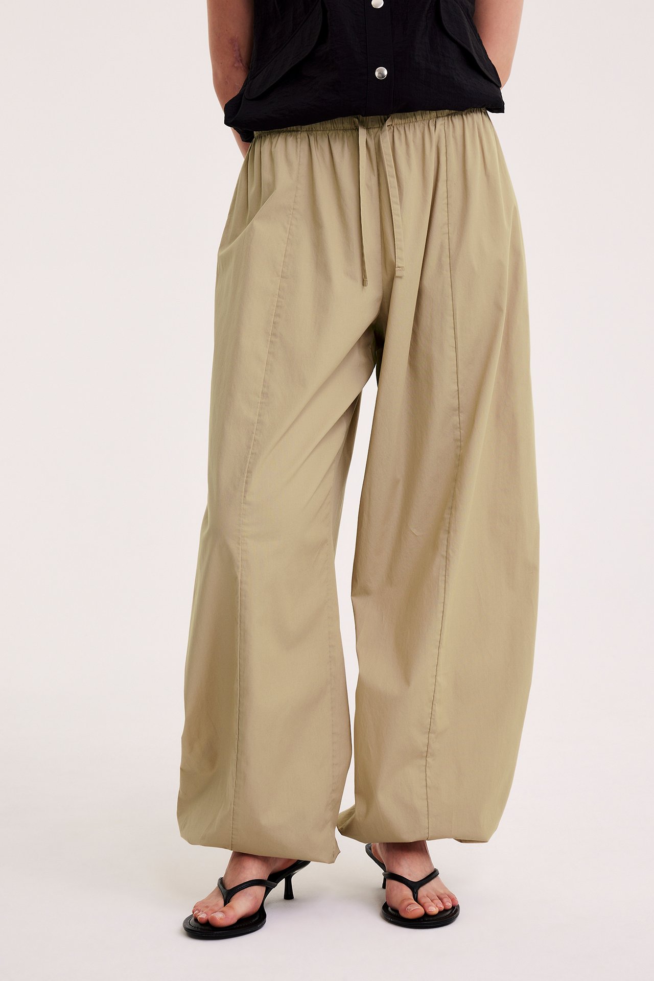 Edestä: Nainen yllään beiget puuvillapopliiniset balloon-housut joustavalla vyötäröllä, musta toppi ja mustat sandaalit.