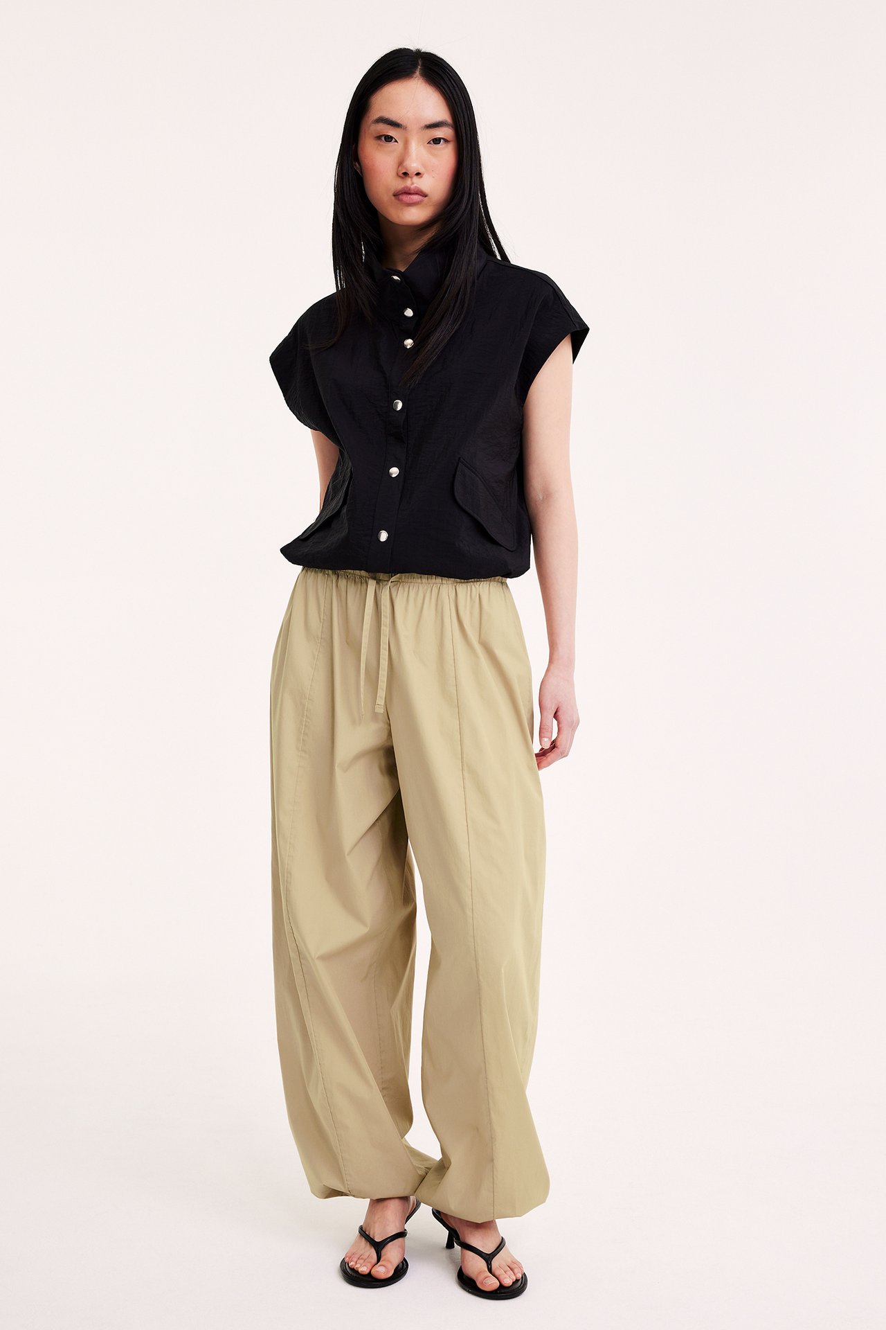 Edestäpäin: nainen beigenvärisissä pallohousuissa, musta hihaton paita ja mustat korkeakorkoiset sandaalit.