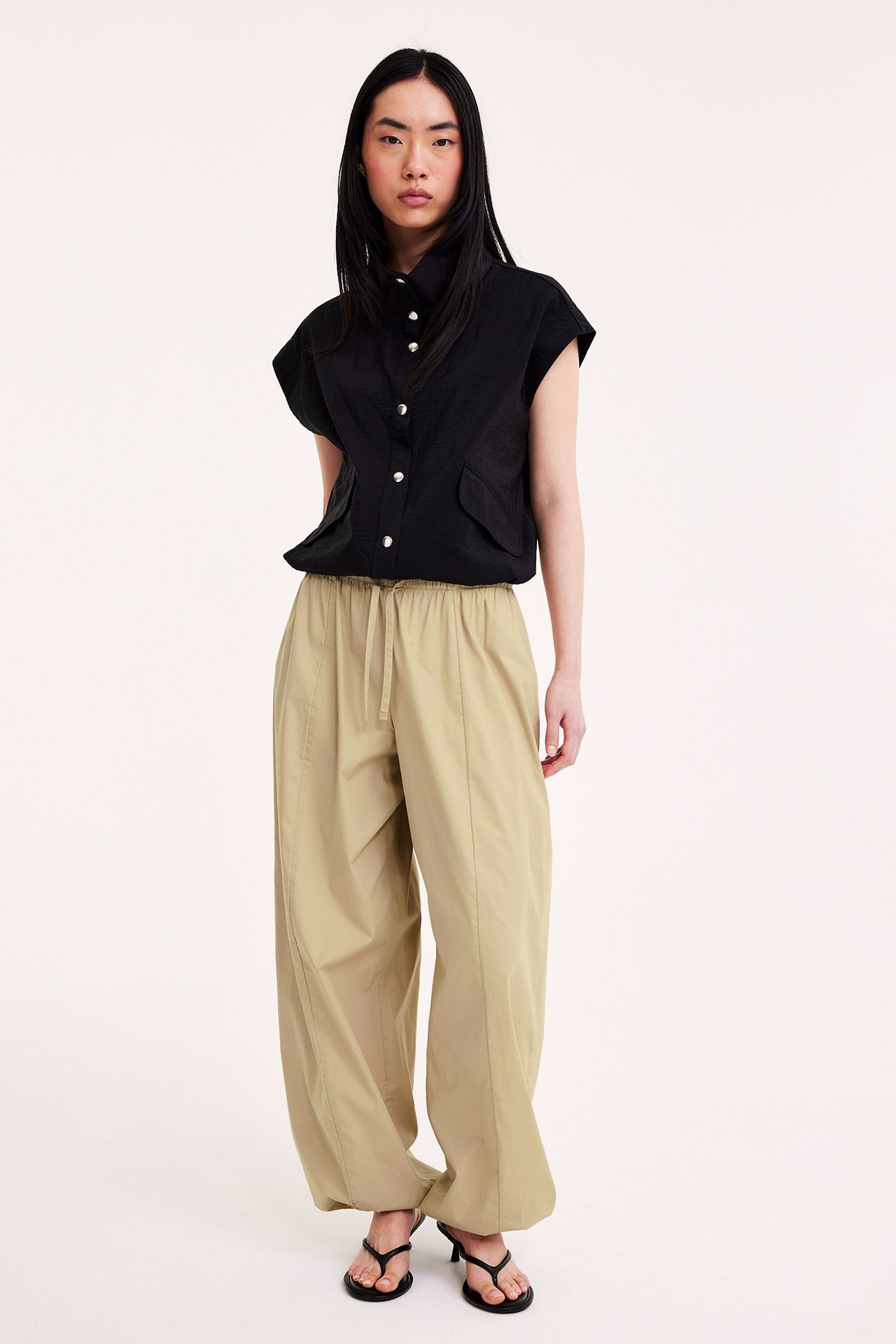 Frontvy av kvinna i beige bomullsballongbyxor, svart ärmlös skjorta och svarta högklackade sandaler.