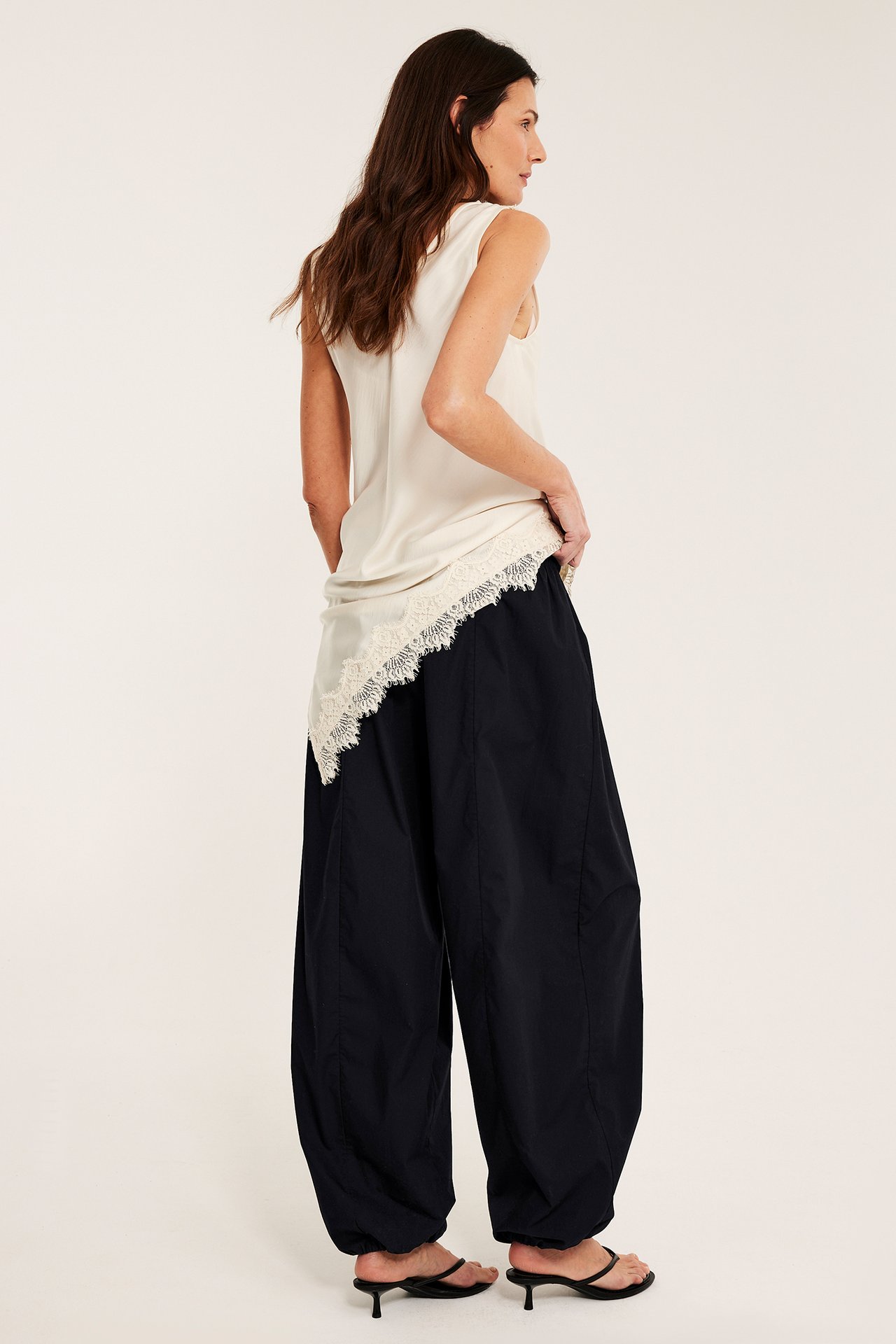 Sida/bakifrån av kvinna i svarta ballongbyxor i bomull, ljusbeige spetstopp, och svarta klacksandaler.