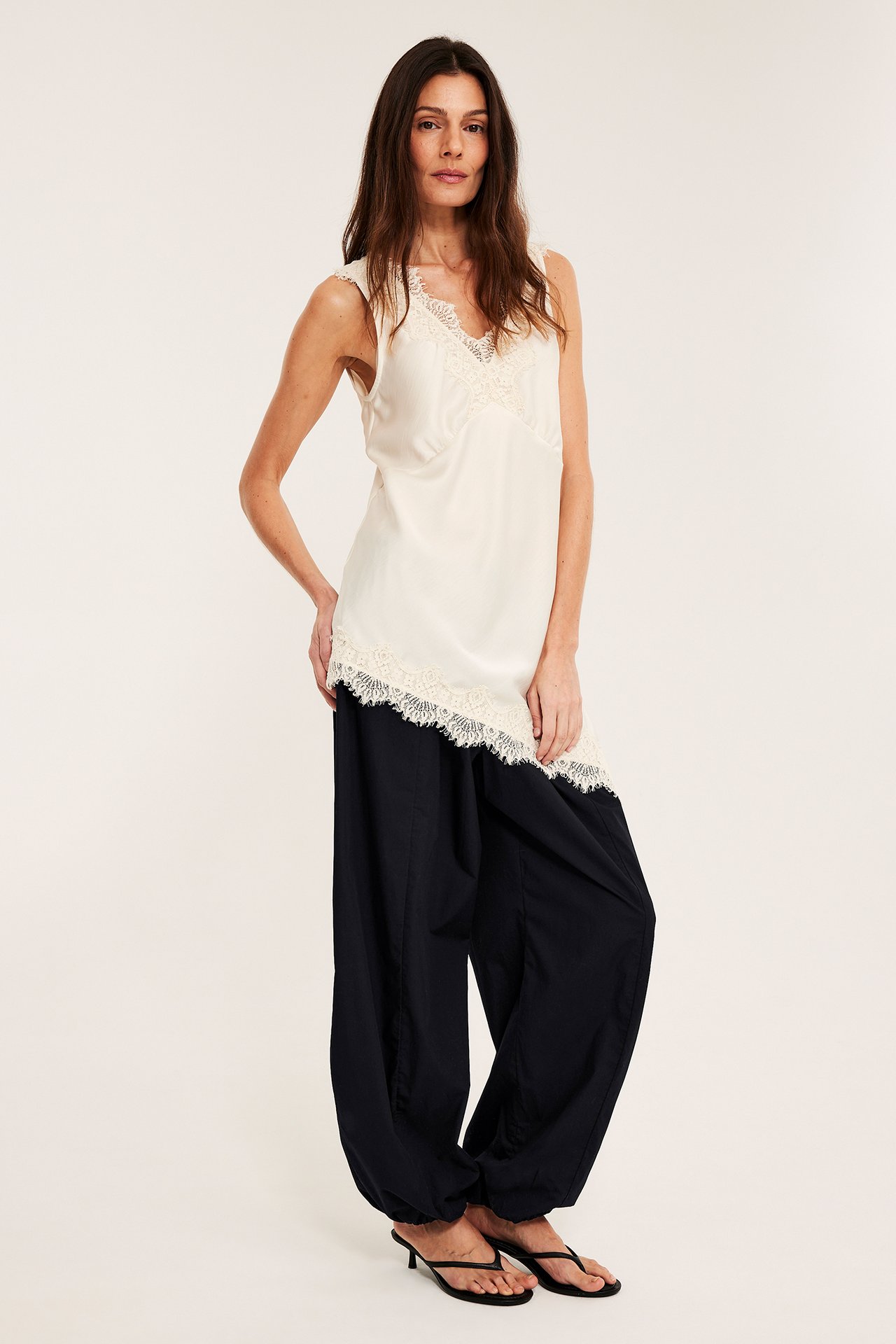 Frontvy av en kvinna i svarta bomullsballongbyxor, en krämvit ärmlös topp med spets och svarta klackflipflops.