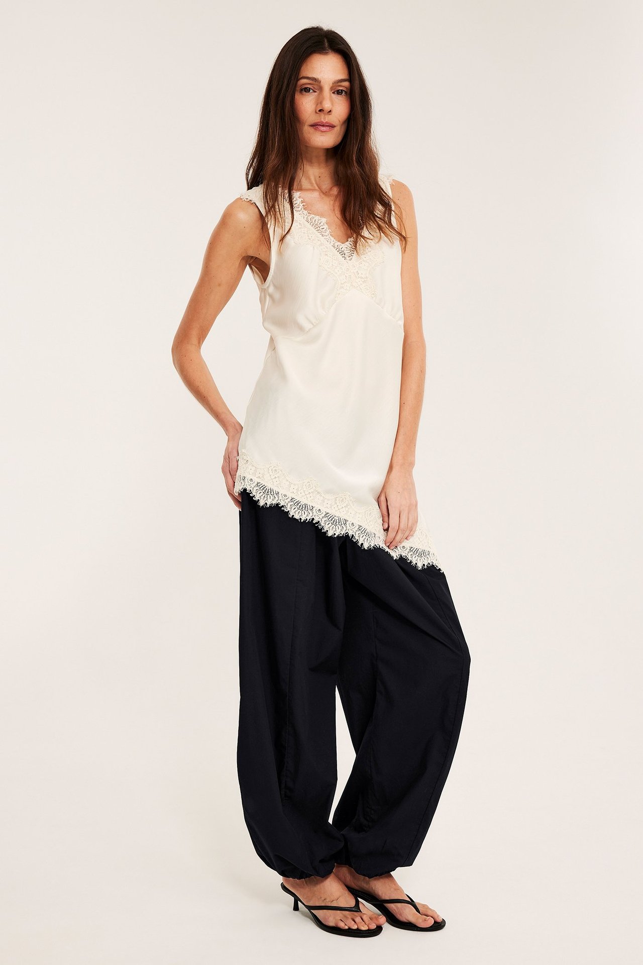 Frontvy av en kvinna i svarta bomullsballongbyxor, en krämvit ärmlös topp med spets och svarta klackflipflops.