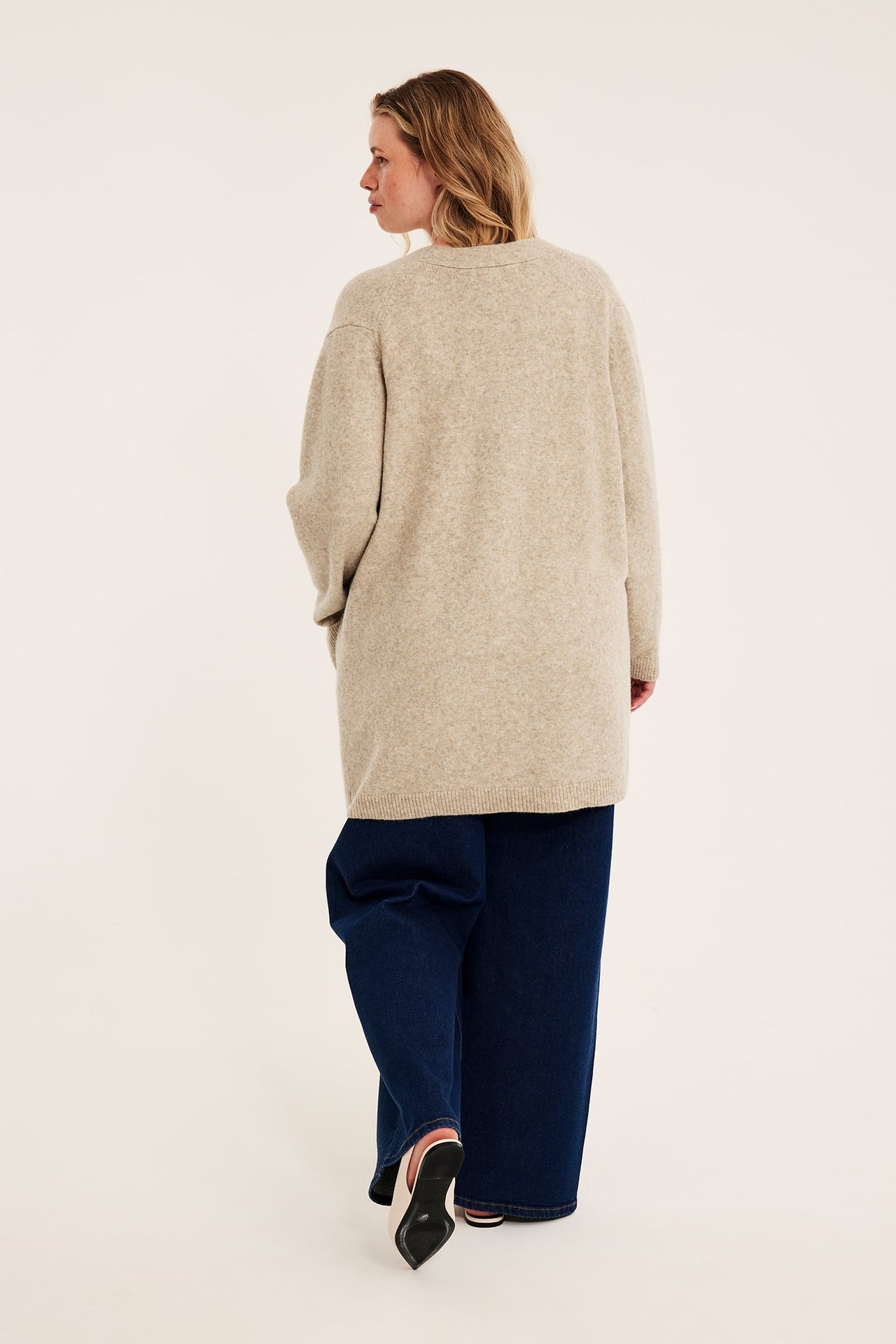 Bakifrån: kvinna i lång beige stickad kofta, mörkblå vida jeans och rosa klackskor.
