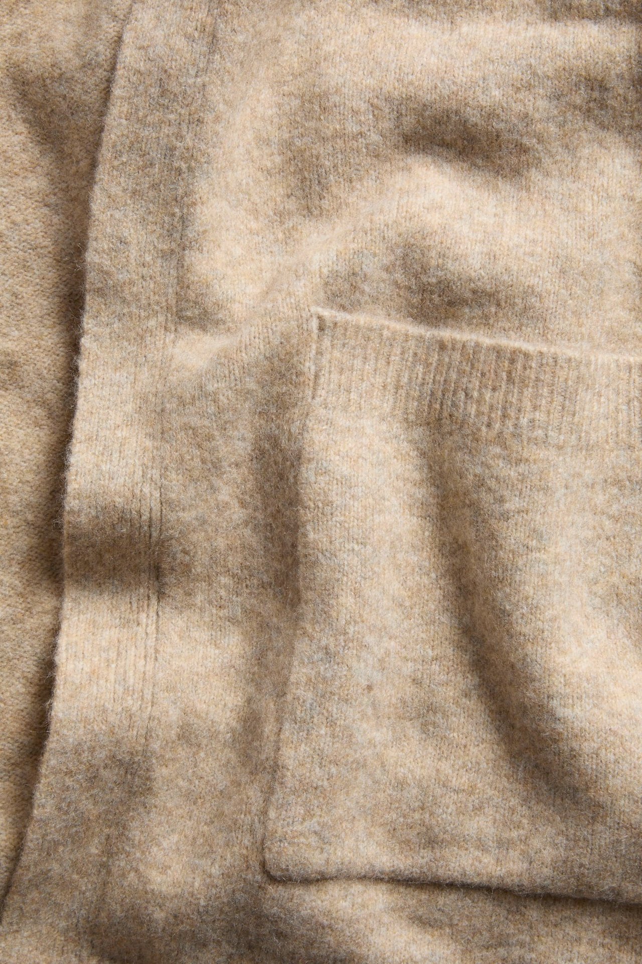 Närbild av en beige melerad lång stickad kofta med en synlig ficka.