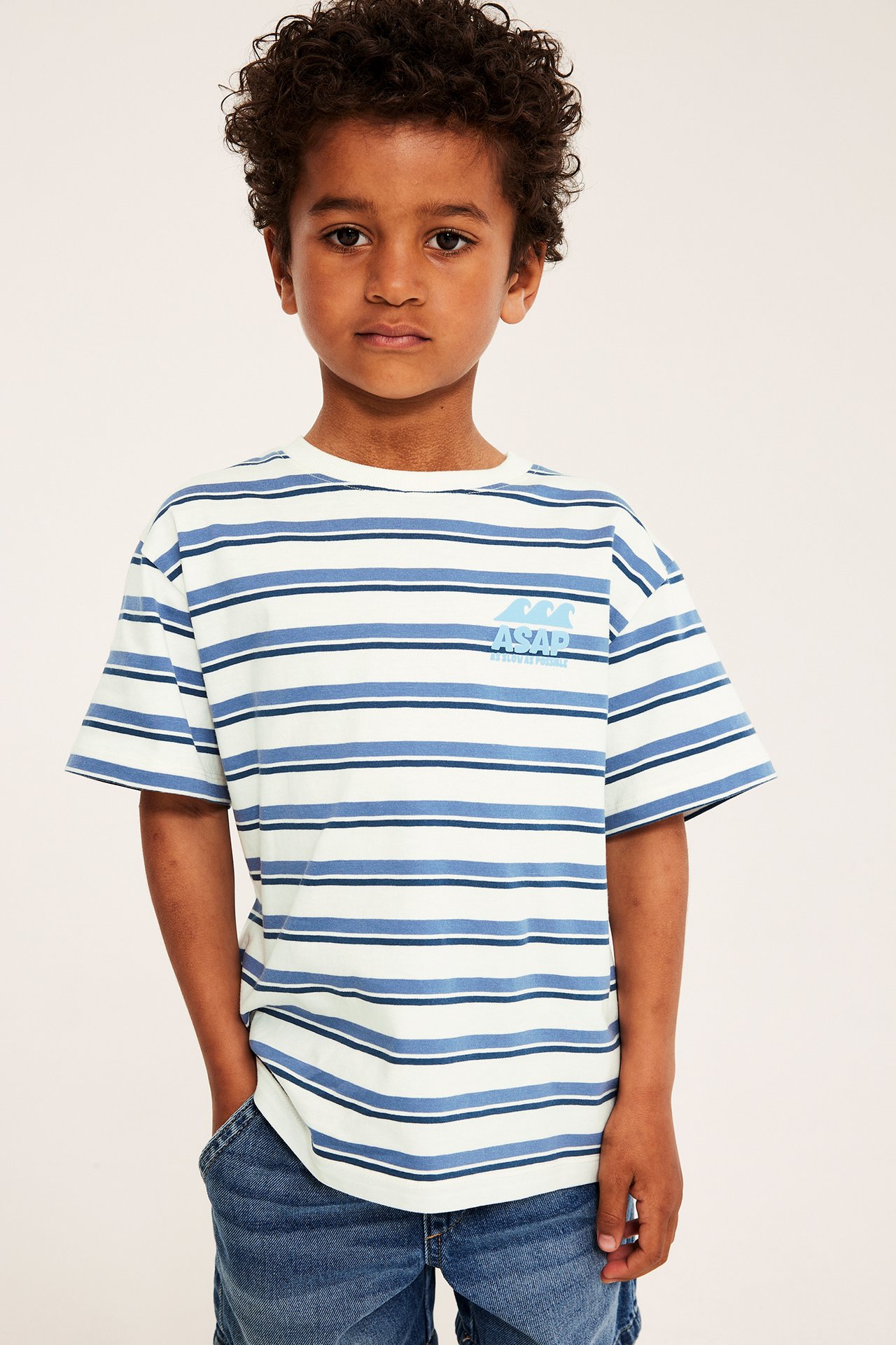 Frontvy av ett barn i en blå- och vitrandig oversize bomulls-t-shirt med broderi, stylad med blå jeans.