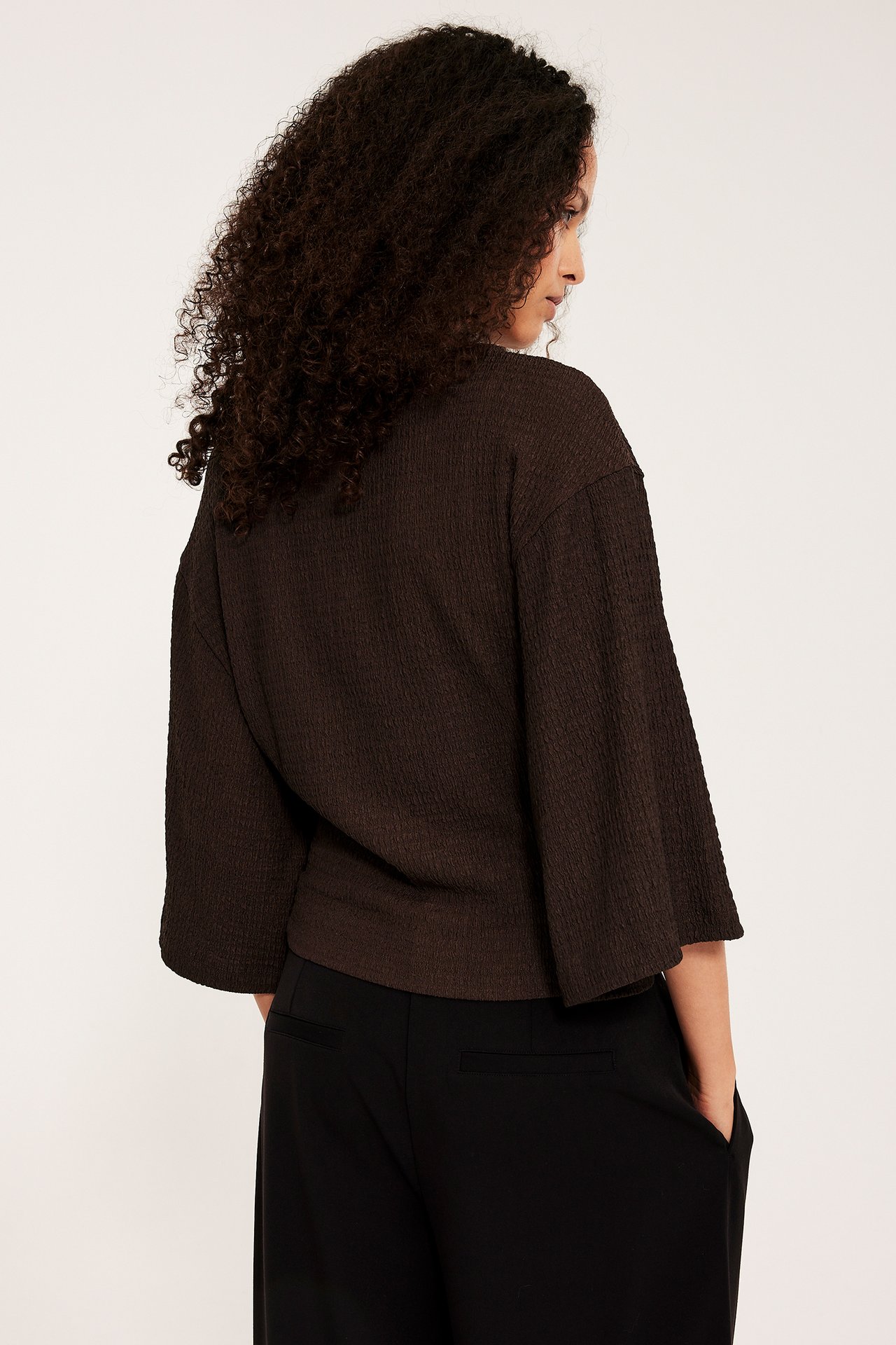 Tummanruskea strukturoitu toppi leveillä kimono-hihoilla naisen päällä, takakuva, stailattu mustilla housuilla.