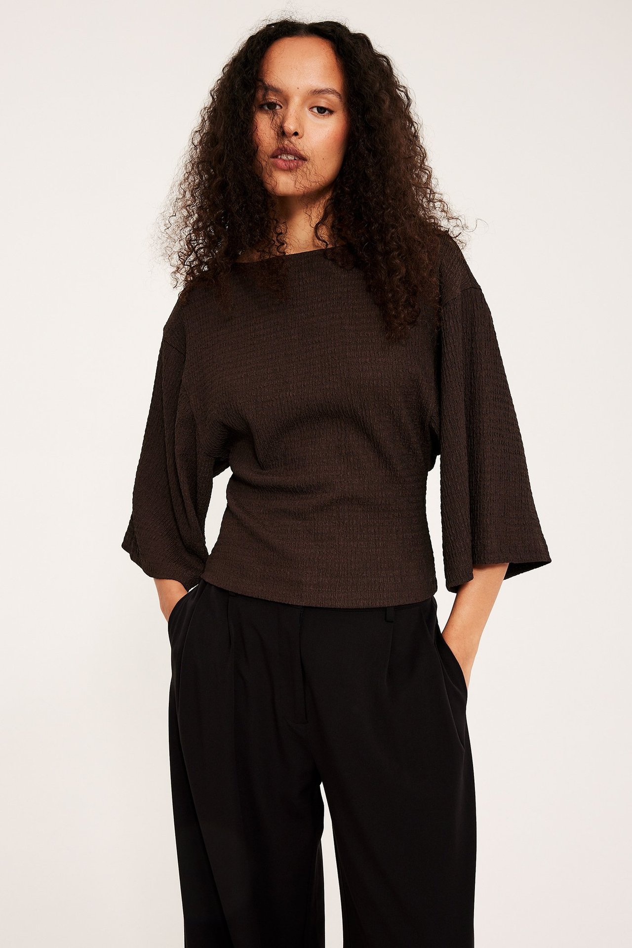 Frontvy av en kvinna i mörkbrun strukturerad topp med vida kimonärmar och svarta byxor.