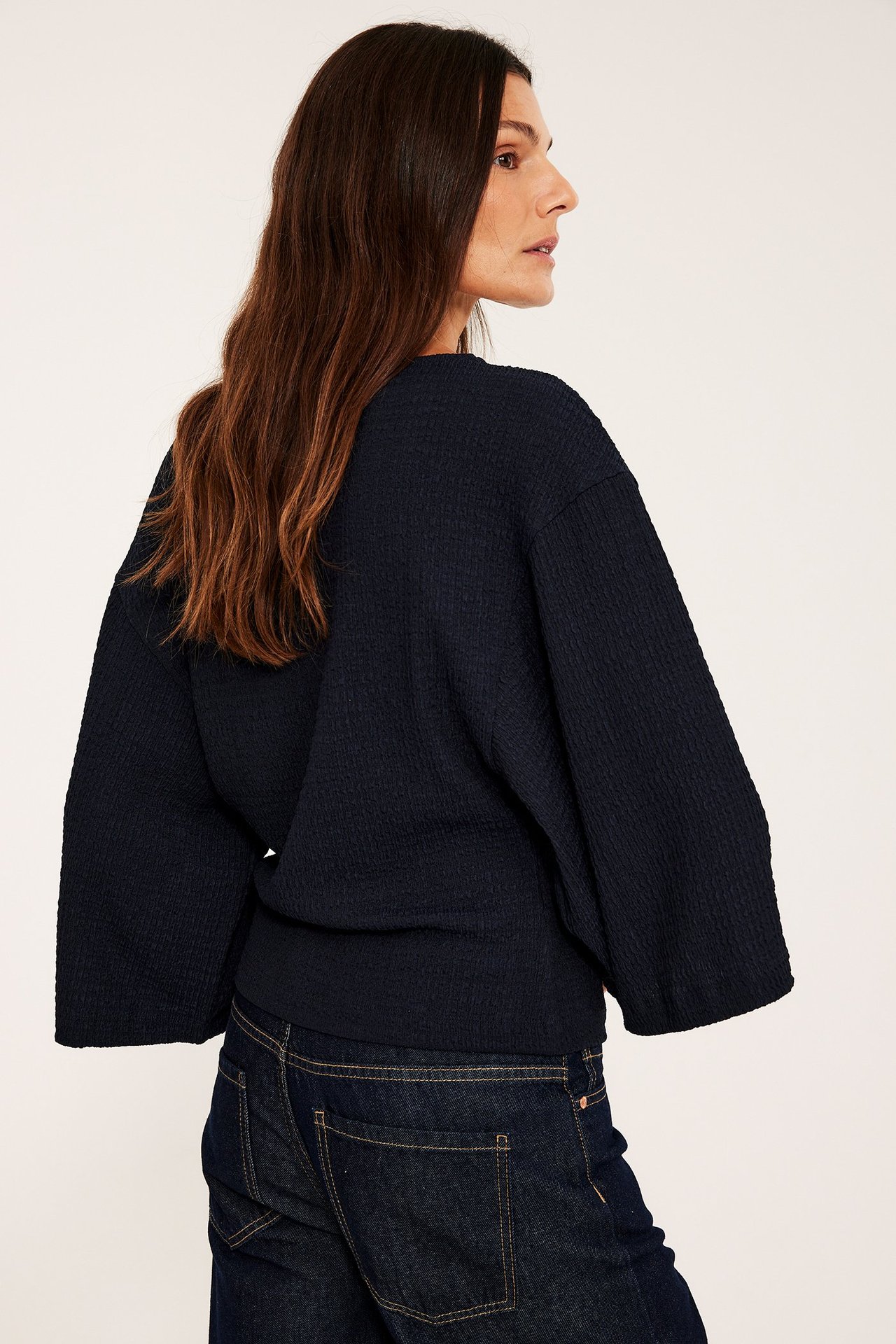 Nainen sivulta/takaa tummansinisessä struktuuritoppissa leveillä kimono-hihoilla ja farkuissa.