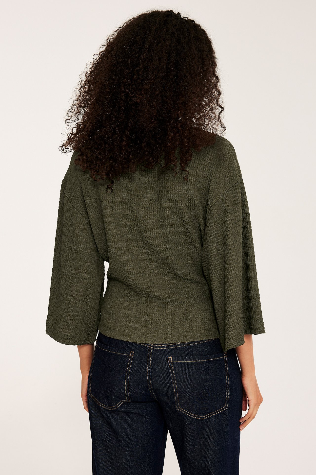 Nainen takaa: vihreä strukturoitu toppi leveillä kimono-hihoilla ja tummansiniset farkut.