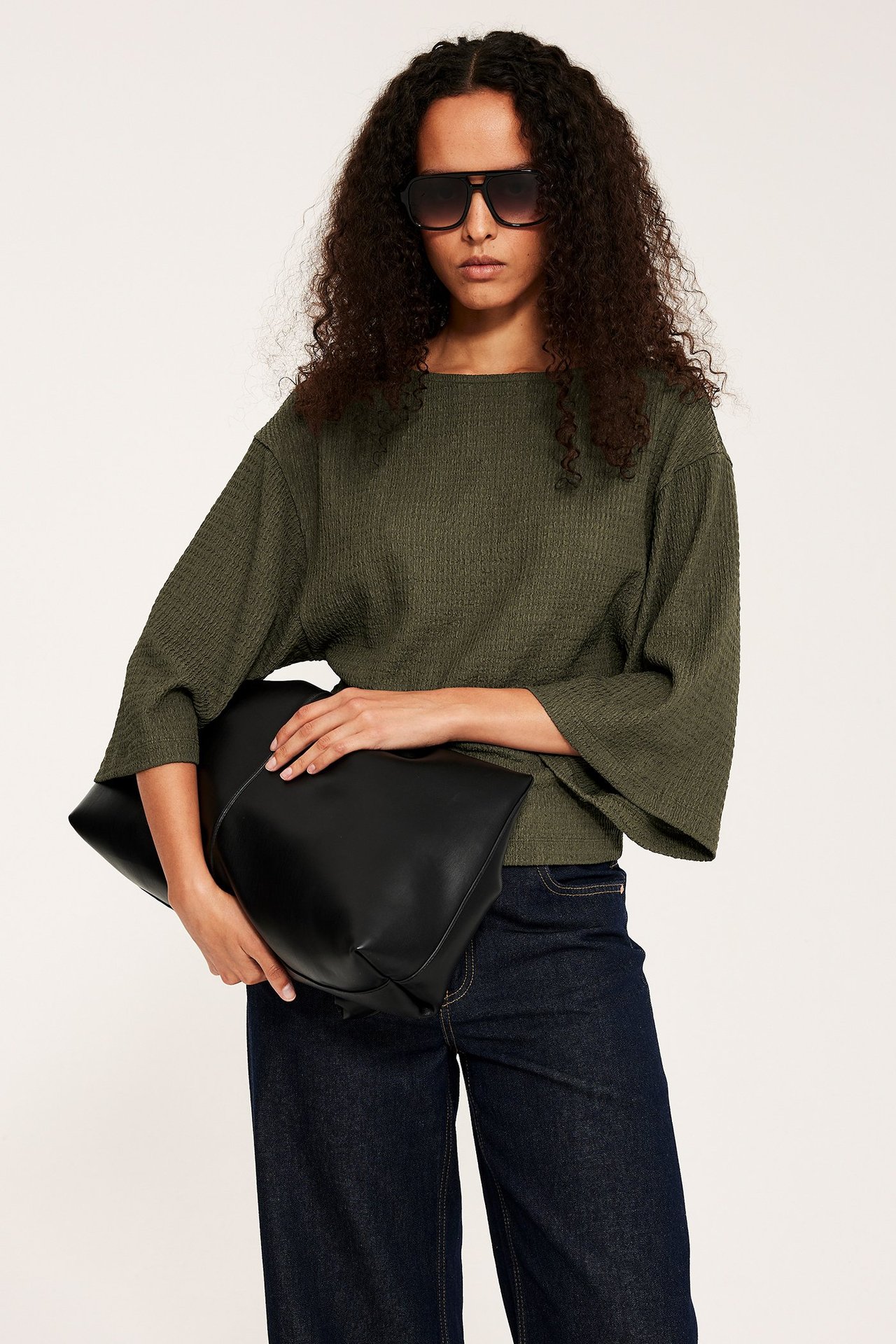 Widok z przodu: kobieta w zielonym strukturalnym topie z szerokimi rękawami, ciemnych dżinsach, okularach, trzymająca czarną torbę.