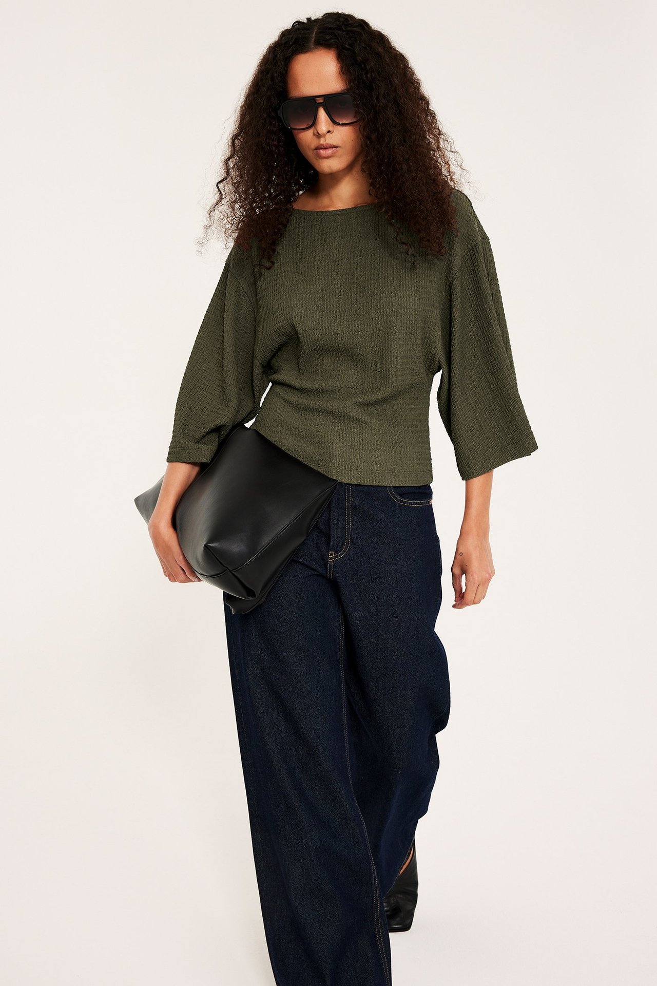 Widok z przodu kobiety w zielonym strukturalnym topie, granatowych jeansach, czarnych okularach, trzymającej czarną kopertówkę.