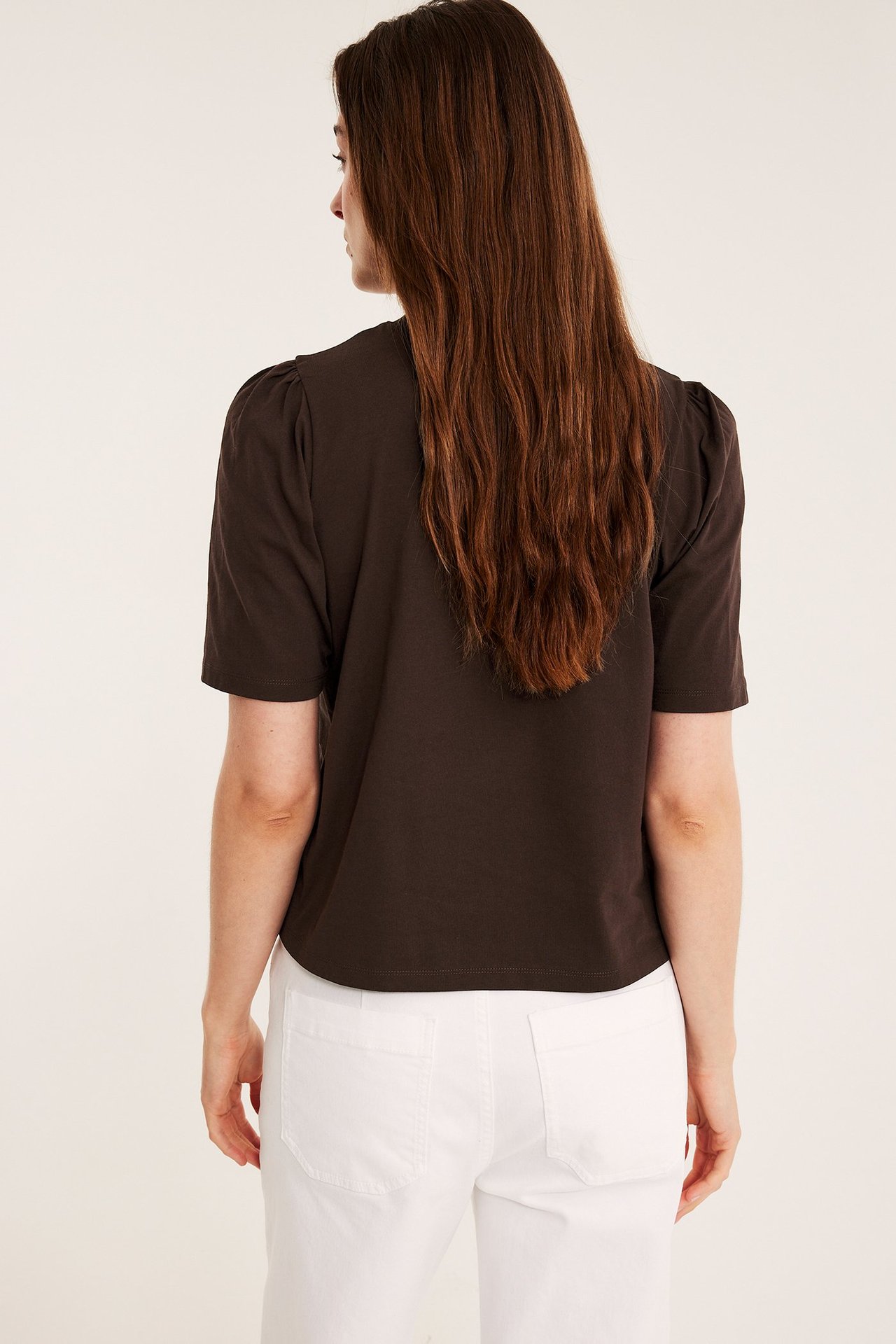 Kobieta tyłem w ciemnobrązowym bawełnianym topie z bufiastymi rękawami i białymi spodniami.