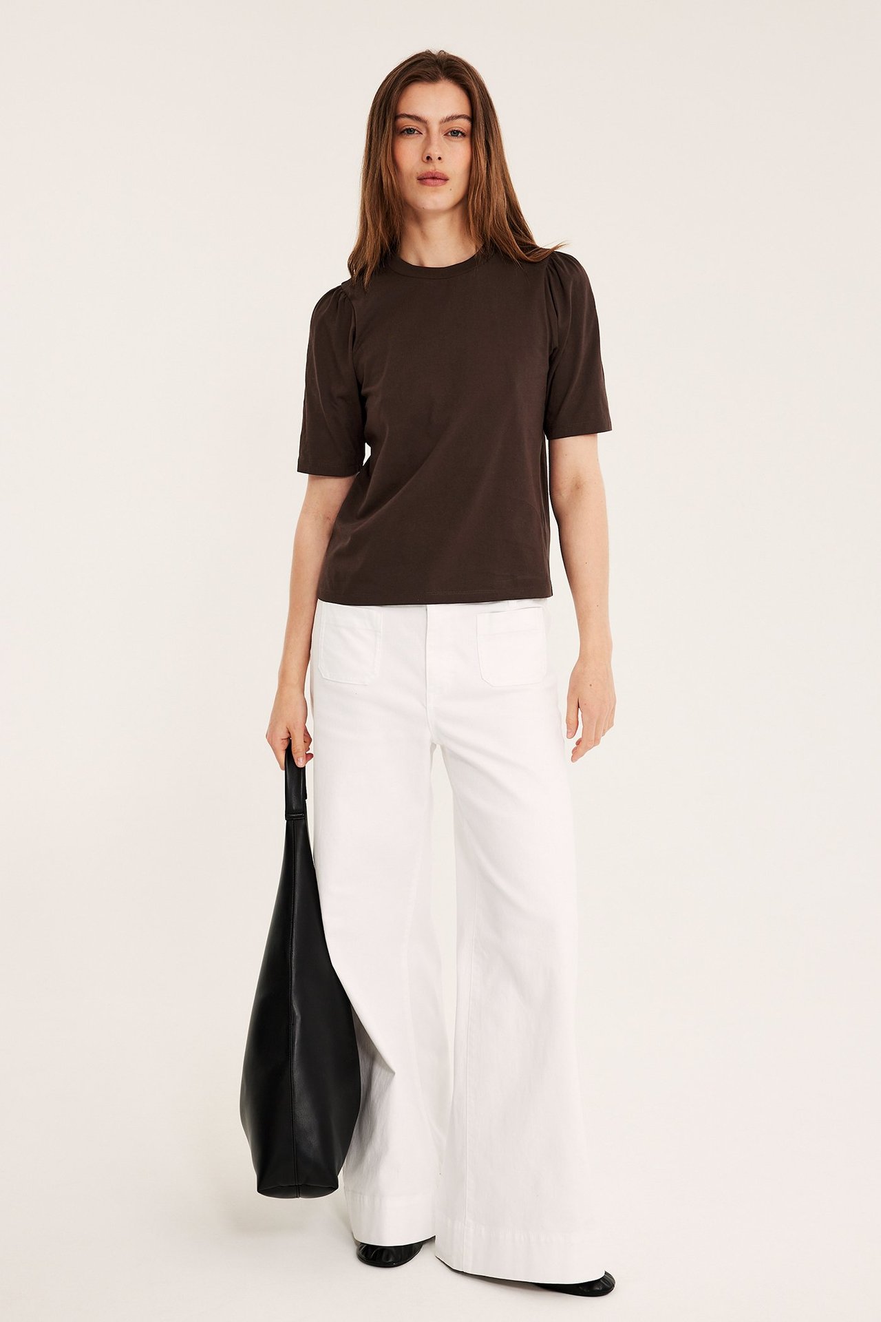 Nainen edestä: tummanruskea puuvillatoppi puffihihoilla, valkoiset leveät housut, musta laukku.