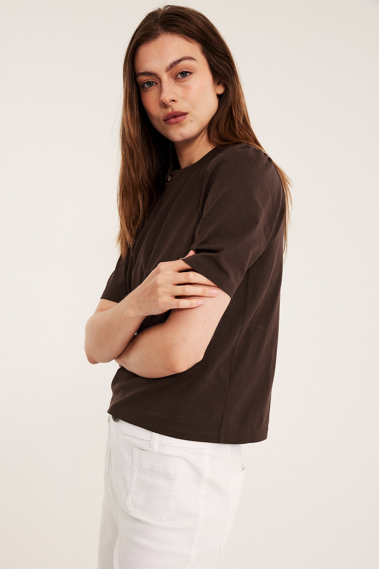 Sivulta edestä näkymä naisesta, jolla on tummanruskea puuvillatoppi pussihihoilla ja valkoiset housut.