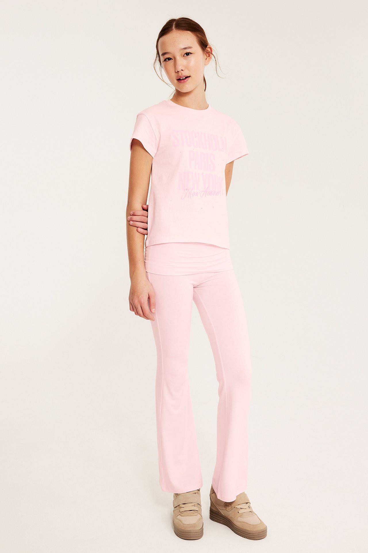 Frontvisning av et barn i lys rosa bomulls-T-skjorte med trykk, lys rosa utsvingte bukser og beige platåsko.