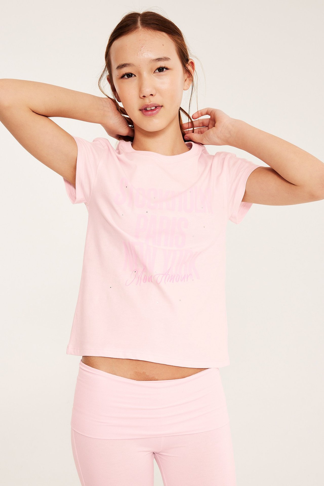 Widok z przodu: dziewczynka ma na sobie jasnoróżowy bawełniany t-shirt z nadrukiem i cyrkoniami, oraz różowe legginsy.