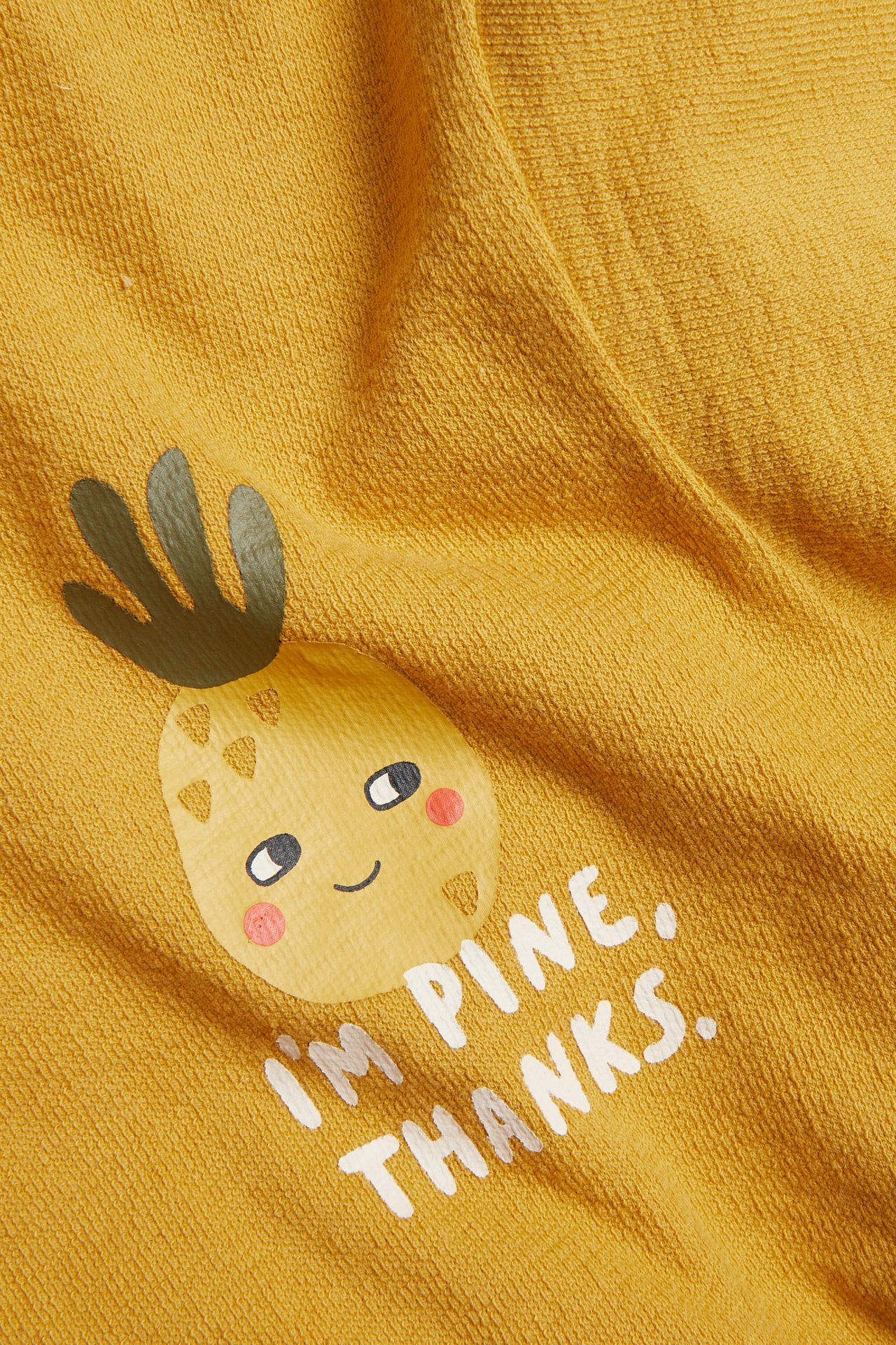 Zbliżenie na żółtą bawełnianą koszulkę dziecięcą z nadrukiem kreskówkowego ananasa i napisem 'I'M PINE. THANKS.'.