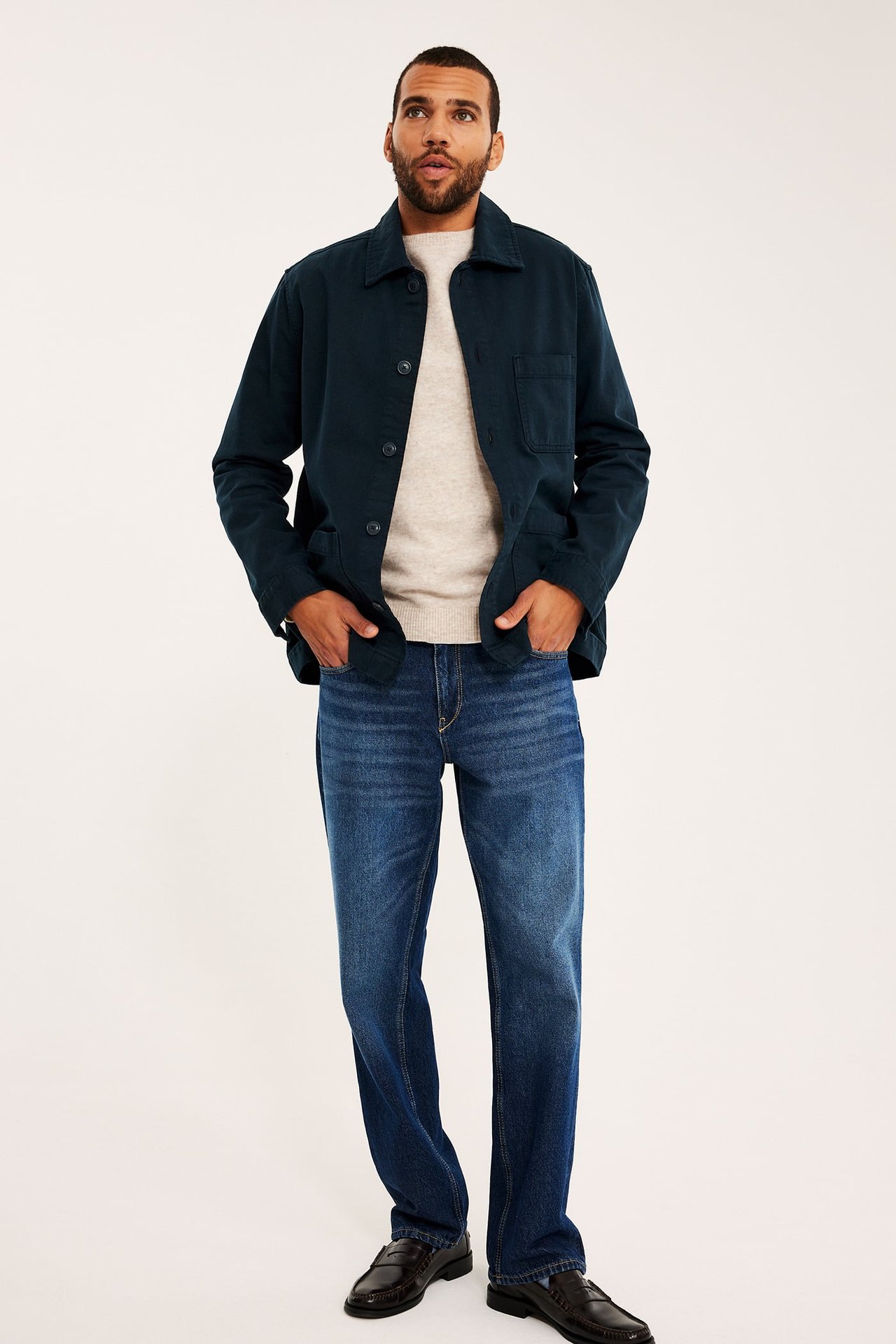 Herr mörkblå workerjacka, ljusbeige tröja, blå jeans och mörkbruna loafers, framifrån.