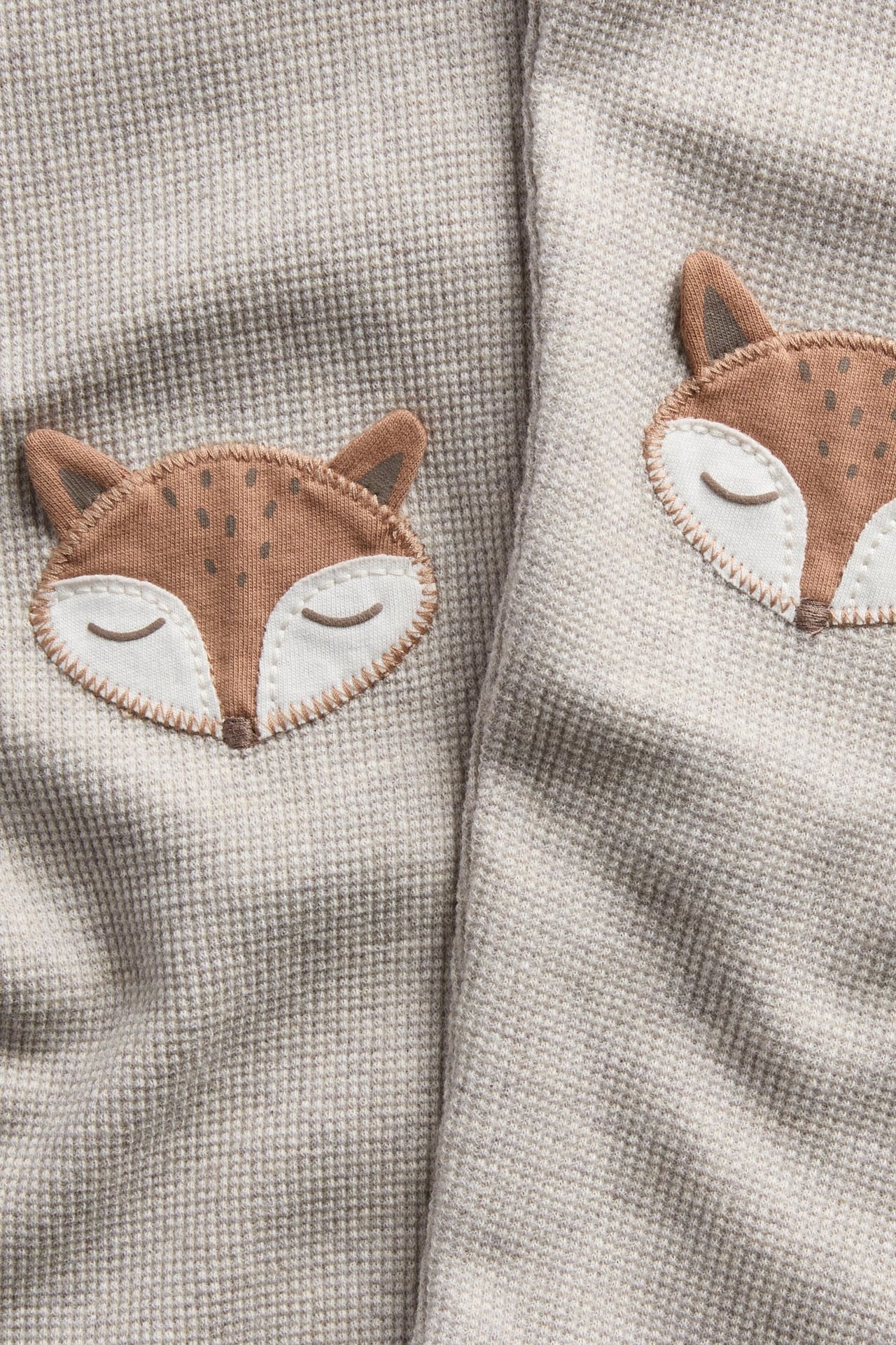Nærbilde av et lys beige vaffelstrikket stoff med to broderte sovende reveapplikasjoner.