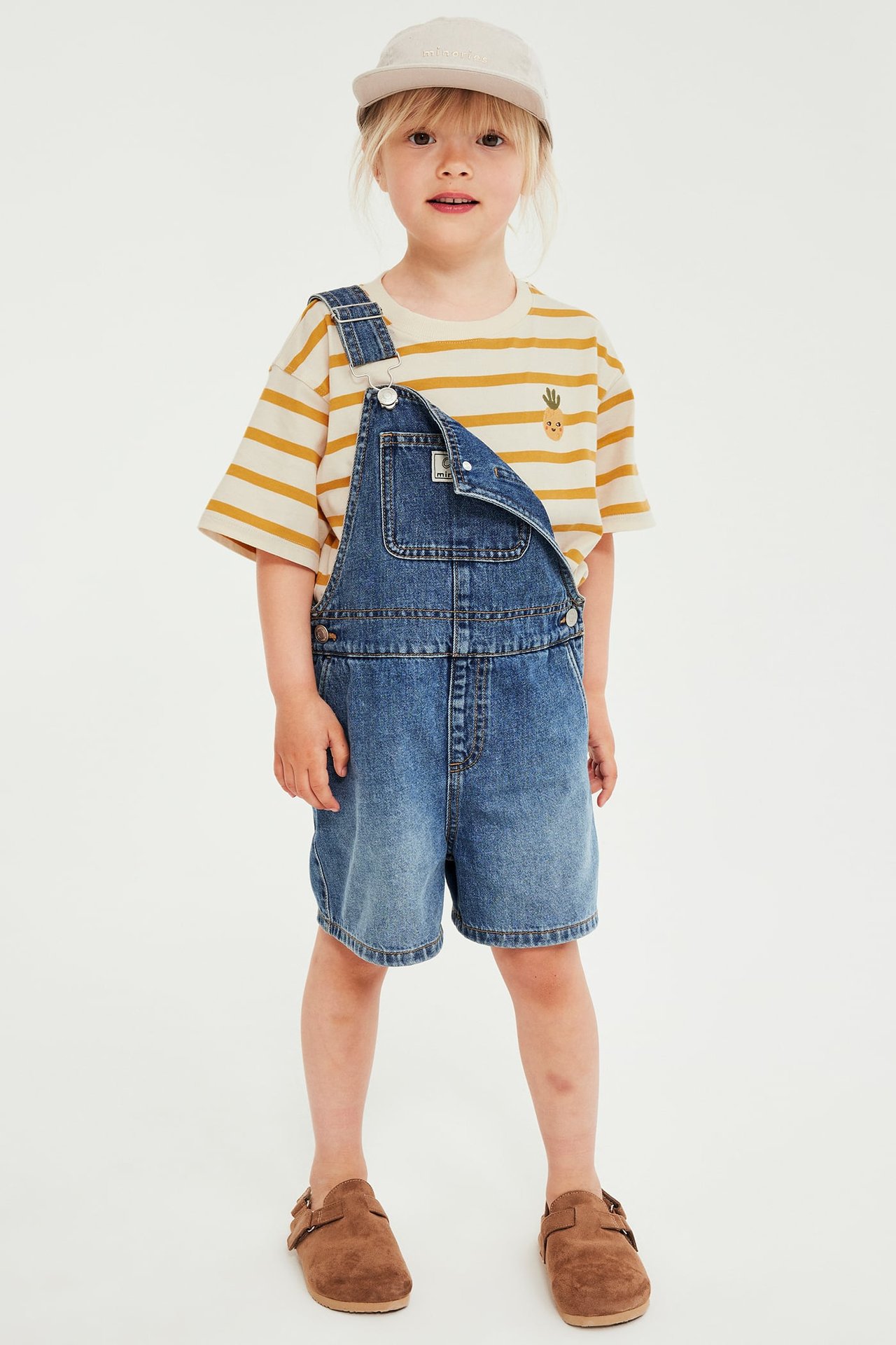Forfra: barn har på seg gulstripet T-skjorte med gulrotbroderi, blå denimshorts-smekkbukse, beige caps, brune tresko.