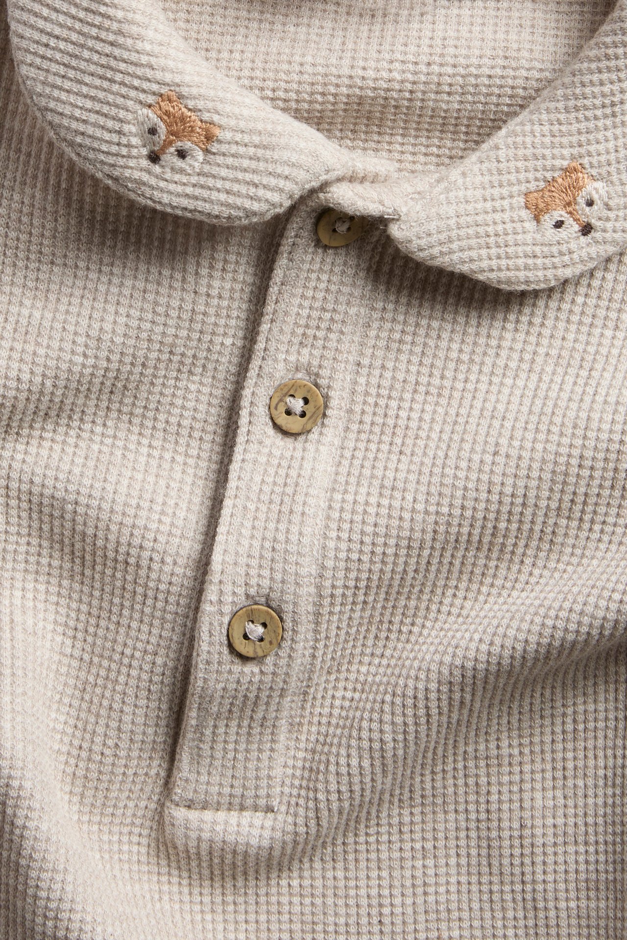 Nærbilde av lys beige vaffelstrikket topp med broderte rever på kragen og to knapper.