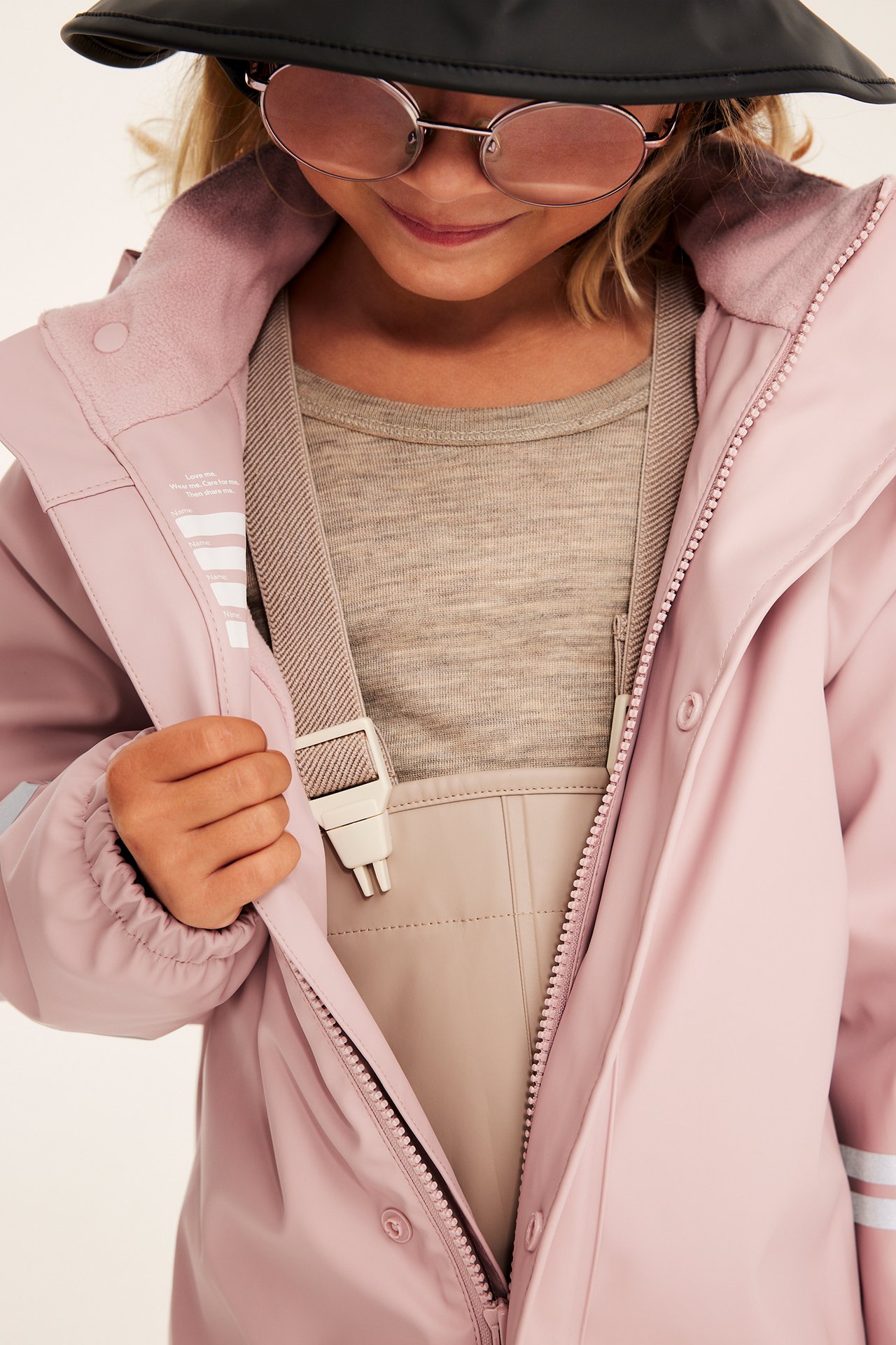 Barn i rosa jakke, åpen over beige snekkerbukse, svart caps og rosa solbriller.