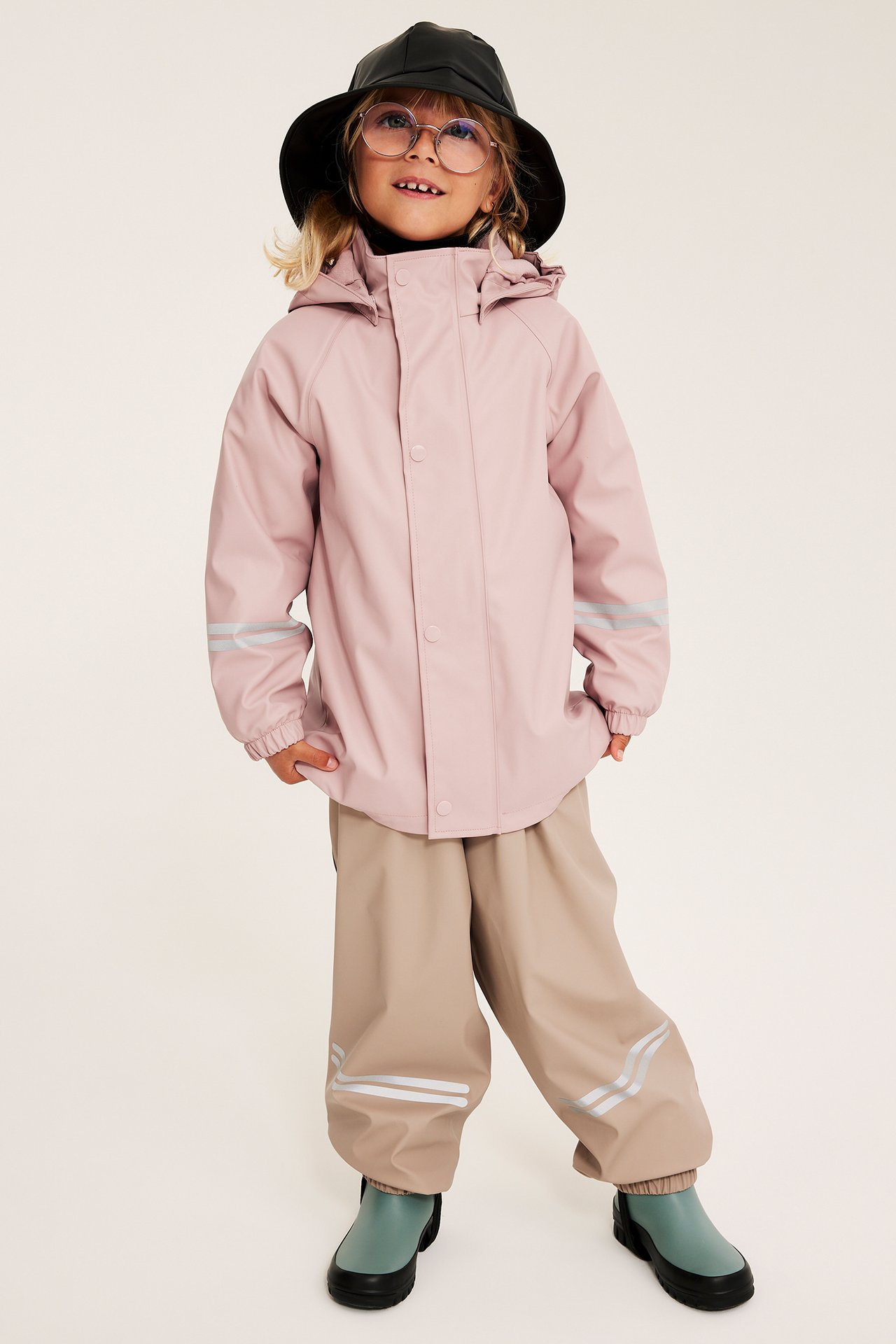 Front barn i rosa regnjakke, beige regnbukser, svart hatt, briller og grønne gummistøvler.