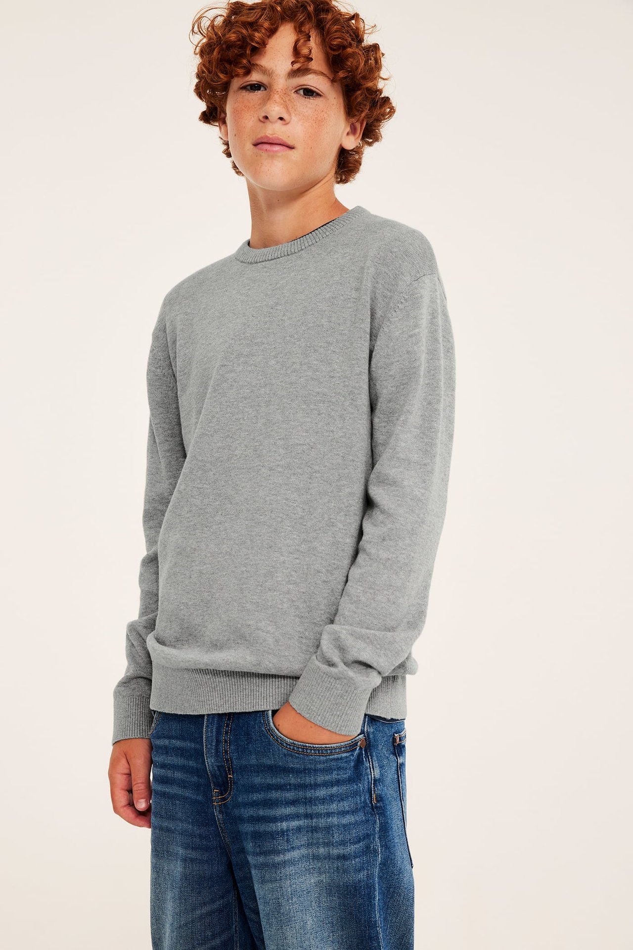 Gutt iført en grå langermet crewneck-genser og blå jeans. Forfra.