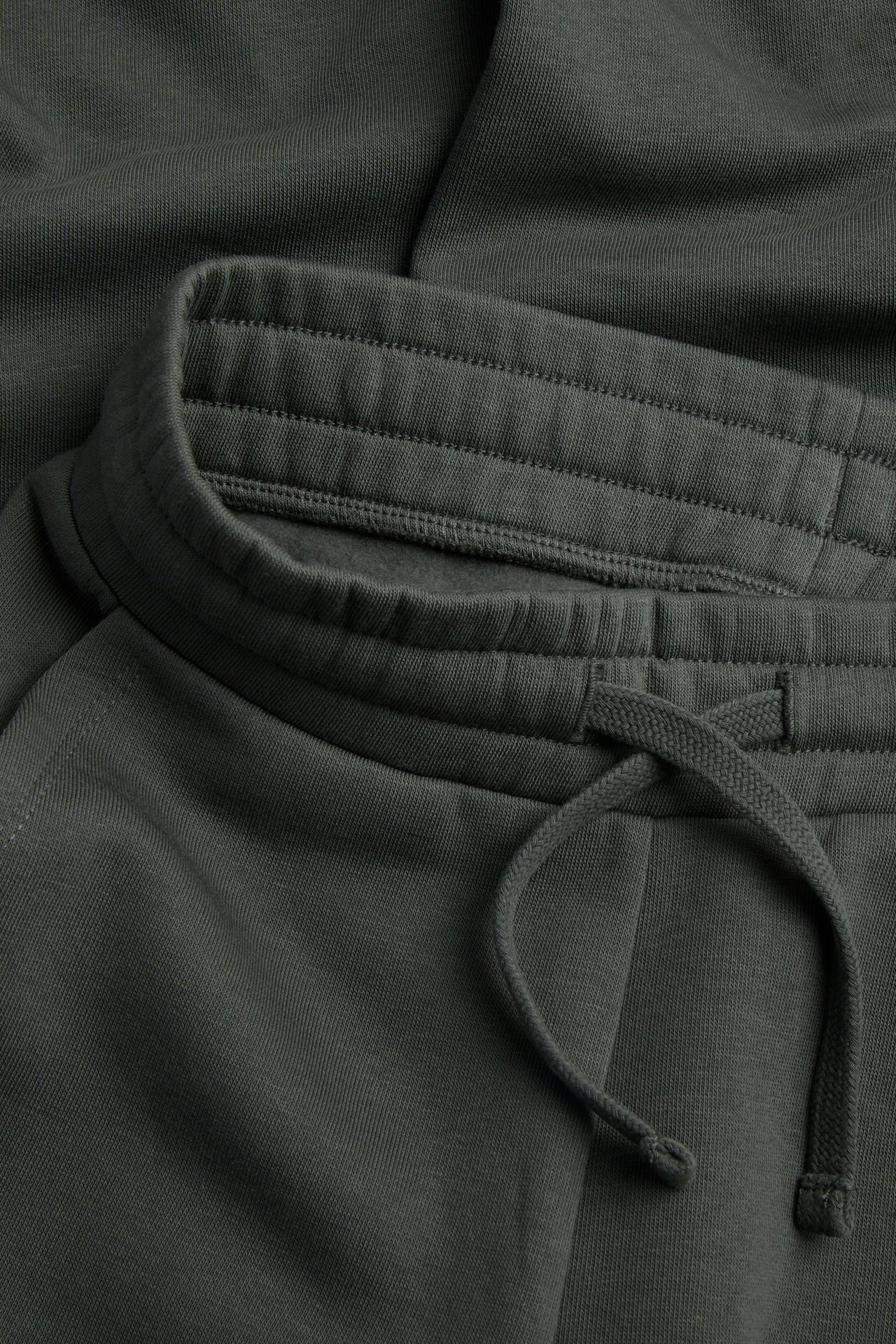 Nærbilde av mørkegrønne ensfargede joggebukser i bomullsblanding med elastisk linning og snøring.