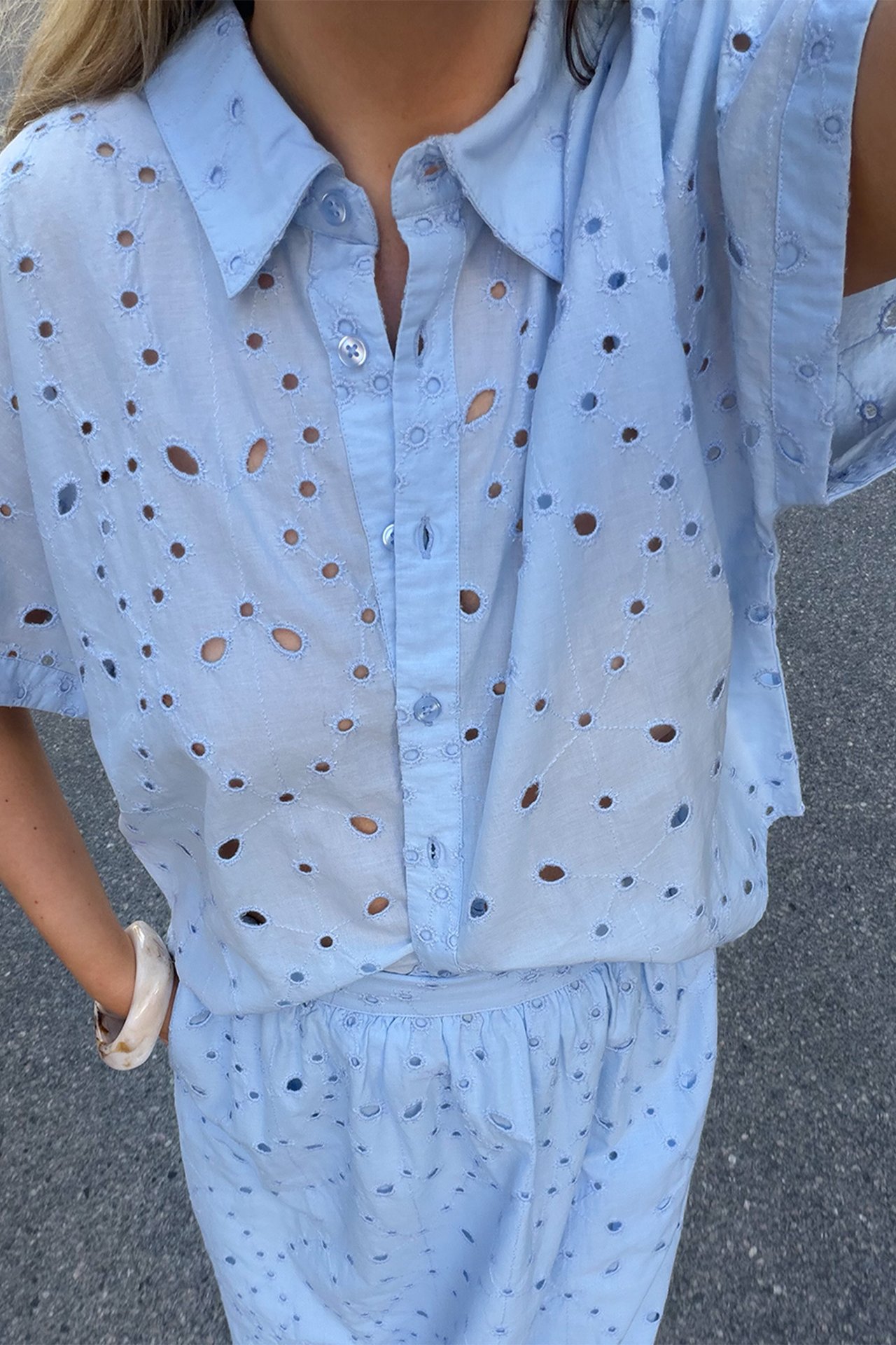 Högvinklad frontvy av en kvinna i ljusblå broderie anglaise bomullsblus, matchande shorts och armring.