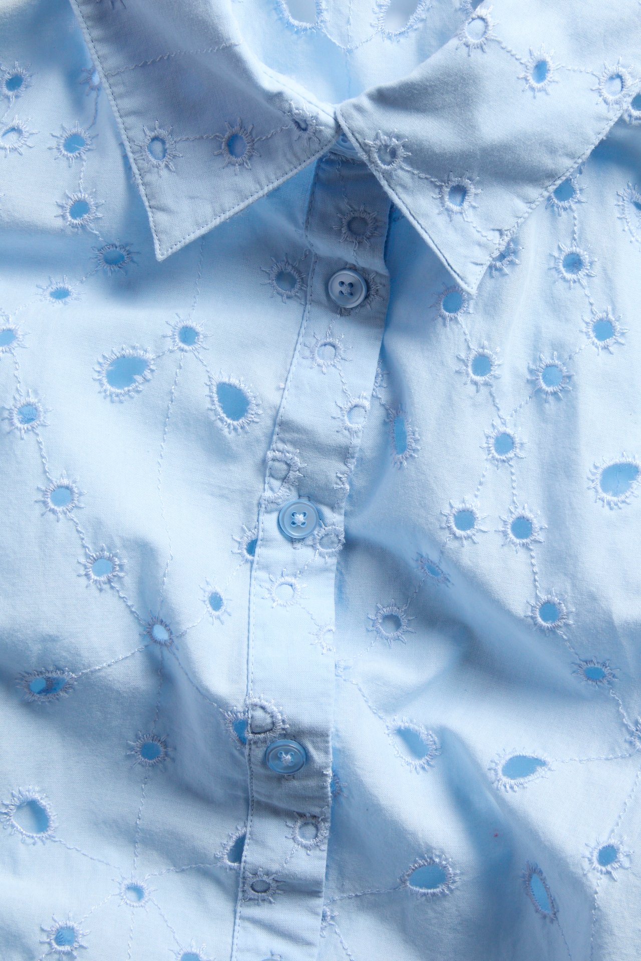 Nærbilde forfra av lys blå bomullsbluse med broderie anglaise-mønster og knapping foran.