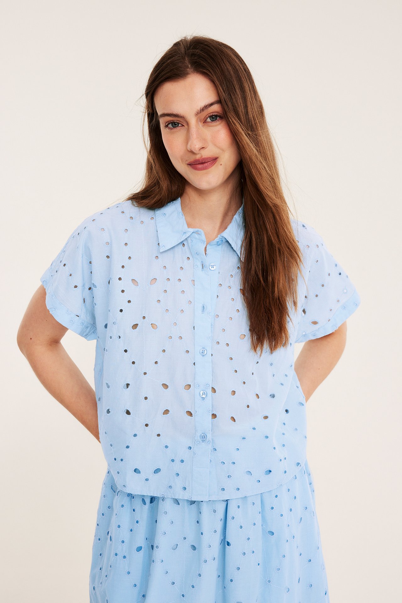 Frontbilde: Kvinne i lysblå kortermet bomullsbluse med broderie anglaise, knapper foran og matchende skjørt.