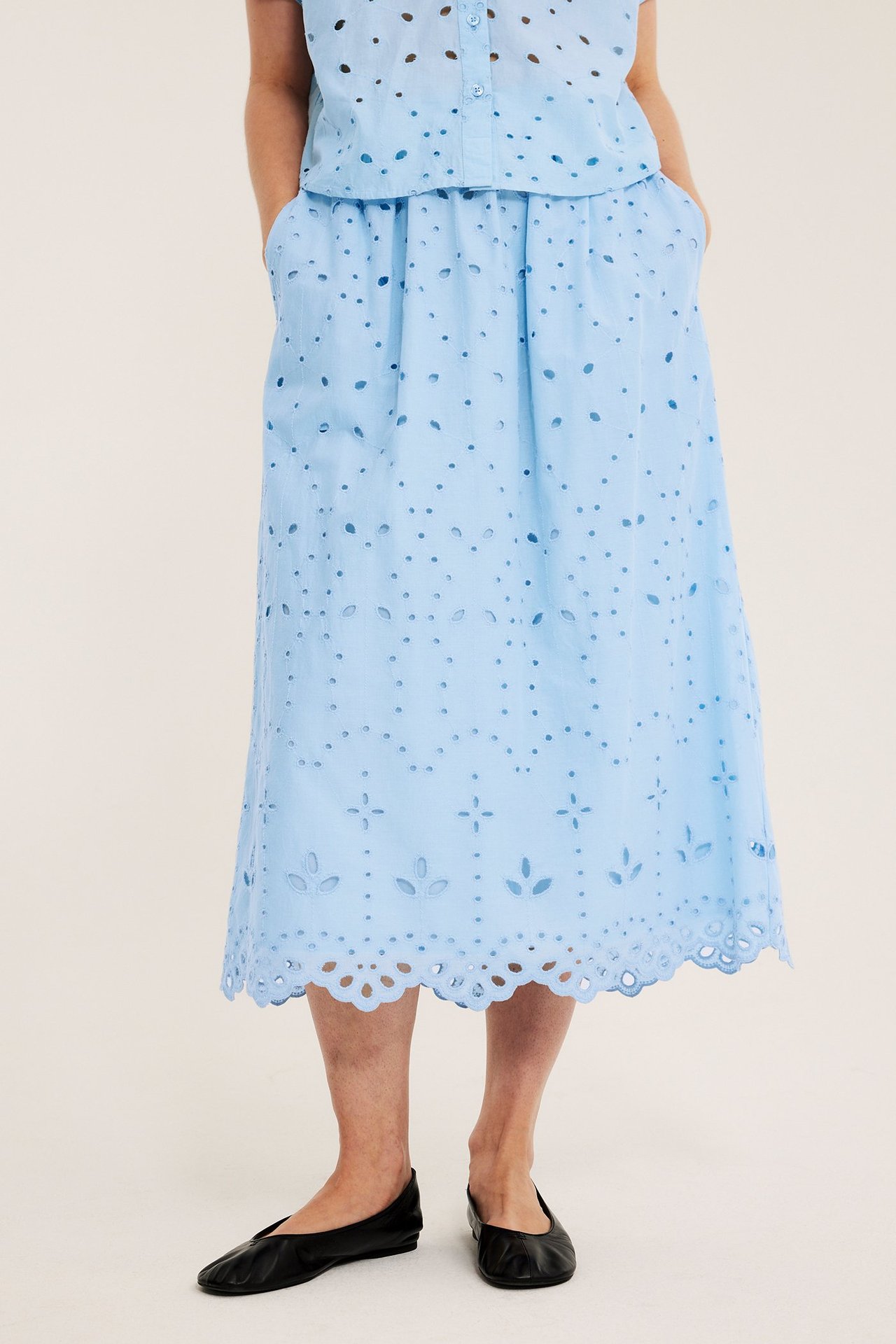 Etunäkymä: Nainen sinisessä broderie anglaise -puuvillahameessa, topissa ja mustissa ballerinakengissä.