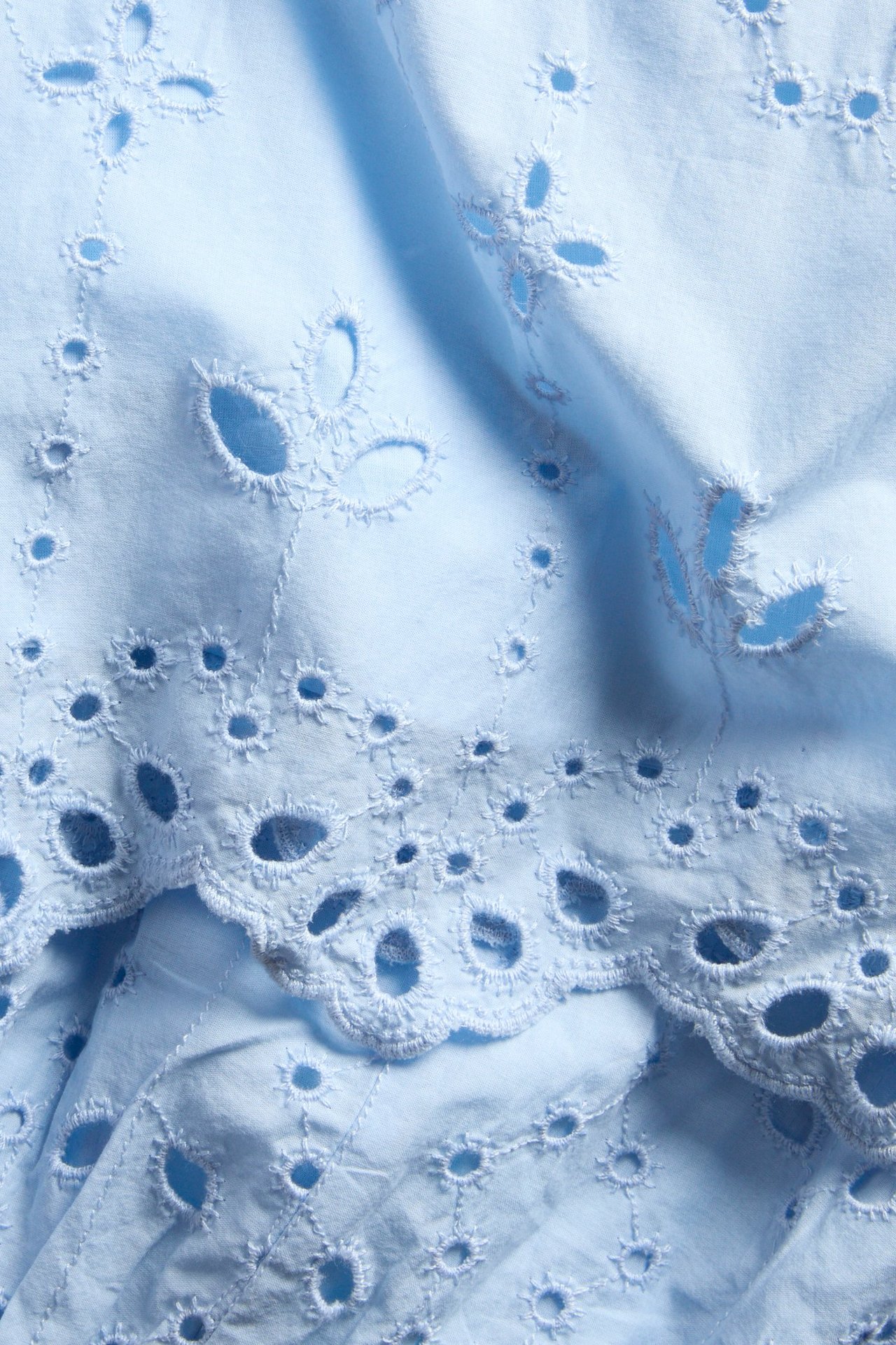 Nærbilde av lyseblått bomullsmidiskjørtestoff med intrikate broderie anglaise utskjærings- og broderidetaljer.