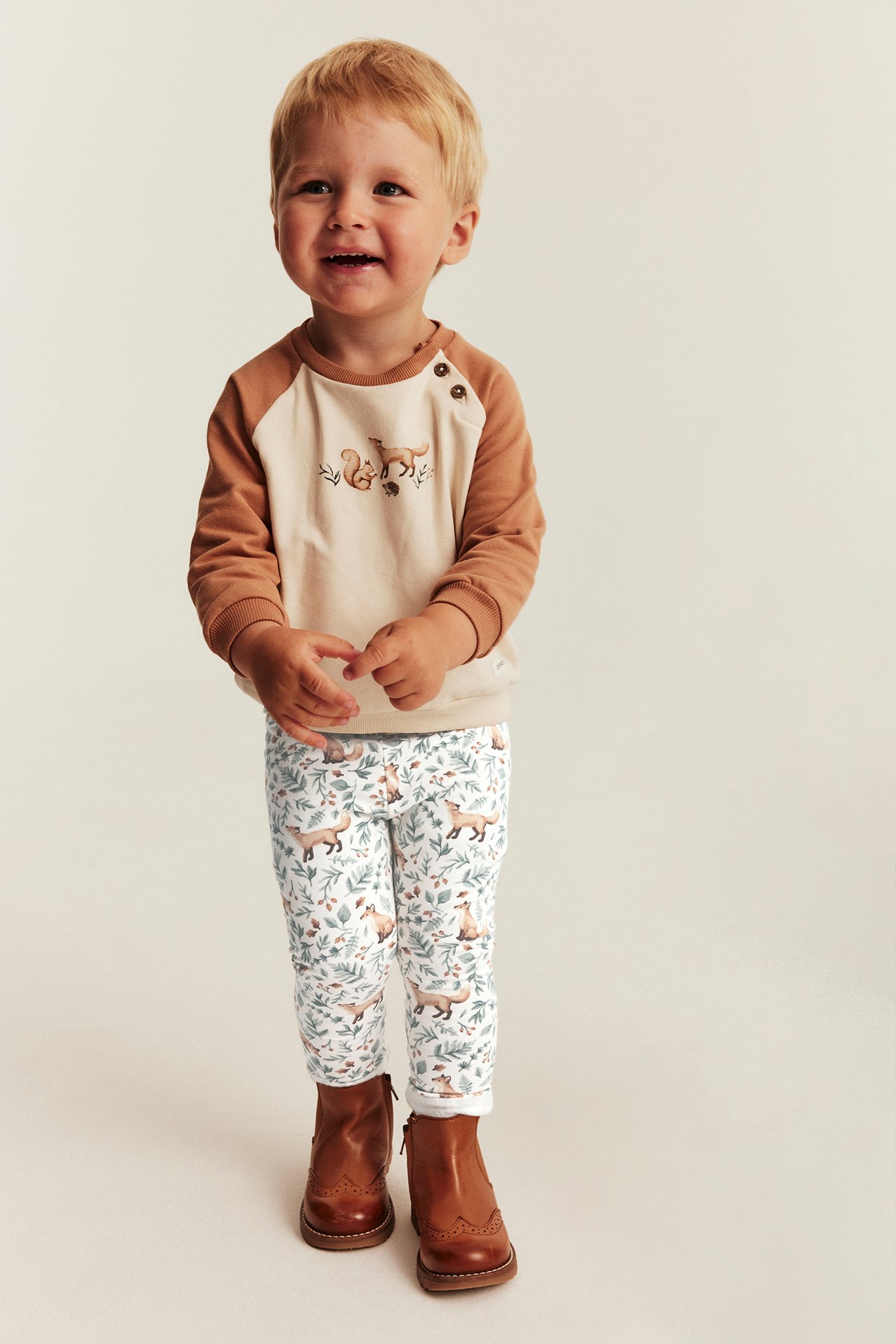 Edestä: lapsi brodeeratussa beige-ruskeassa pitkähihaisessa paidassa, valkoisissa kuviollisissa leggingseissä ja ruskeissa saappaissa.