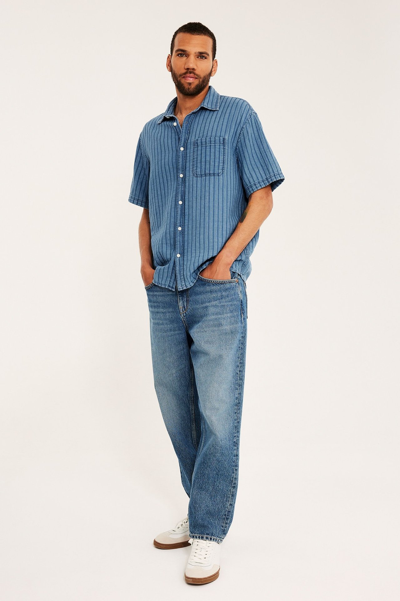 Framifrån, en man i en ljus denimrandig kortärmad skjorta med bröstficka, blå jeans och vita sneakers.