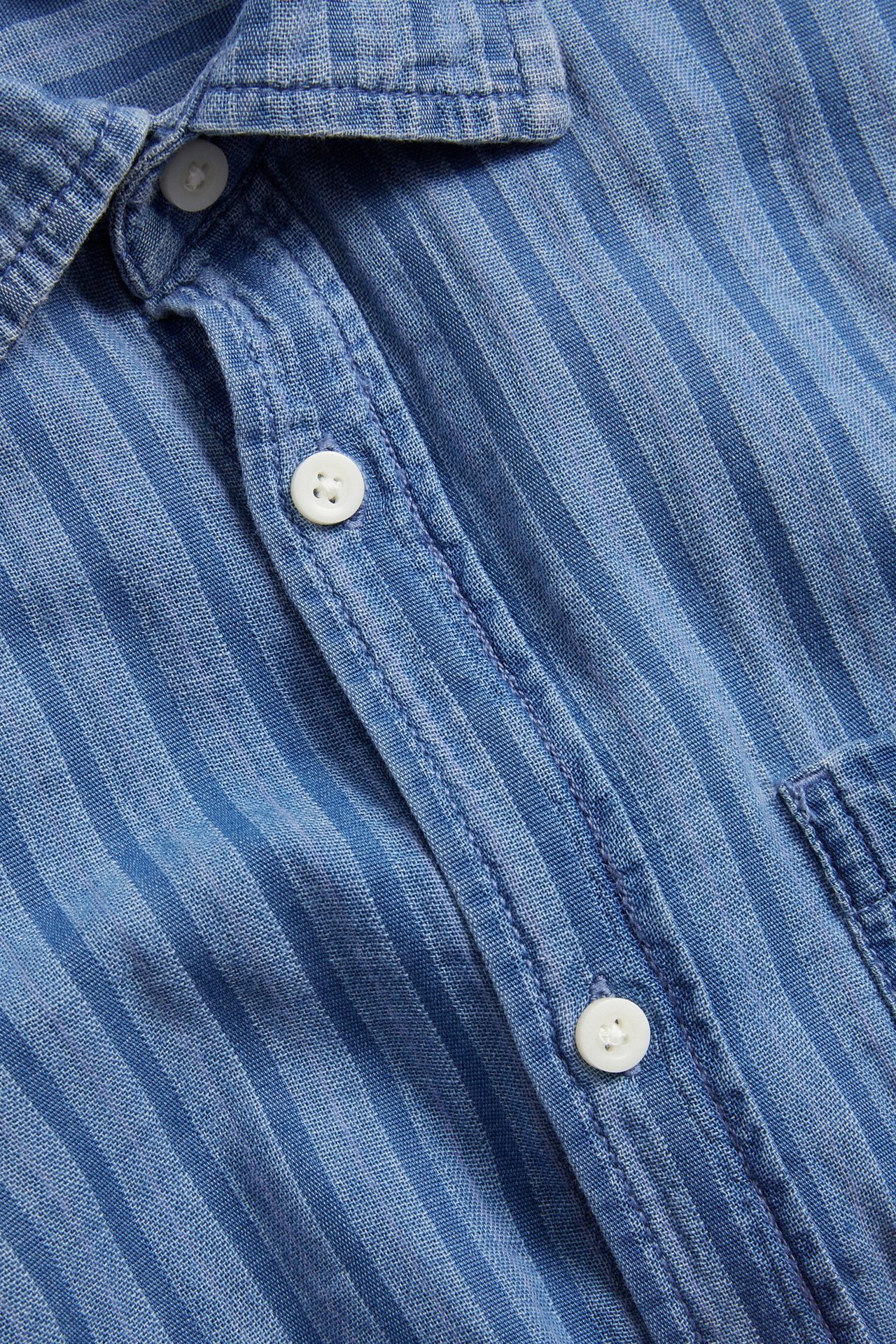 Närbild av en herrskjorta i ljusblå randig denim med normal krage, vita knappar och bröstficka.