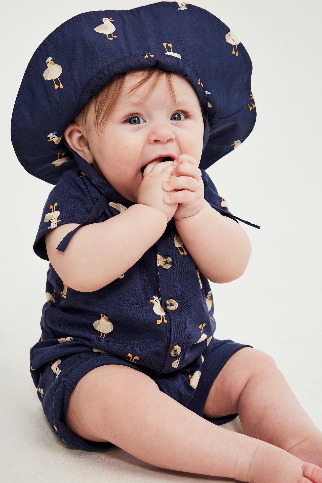 Framifrån: en baby i mörkblå kortärmad romper i bomull med ankmönster och matchande hatt.