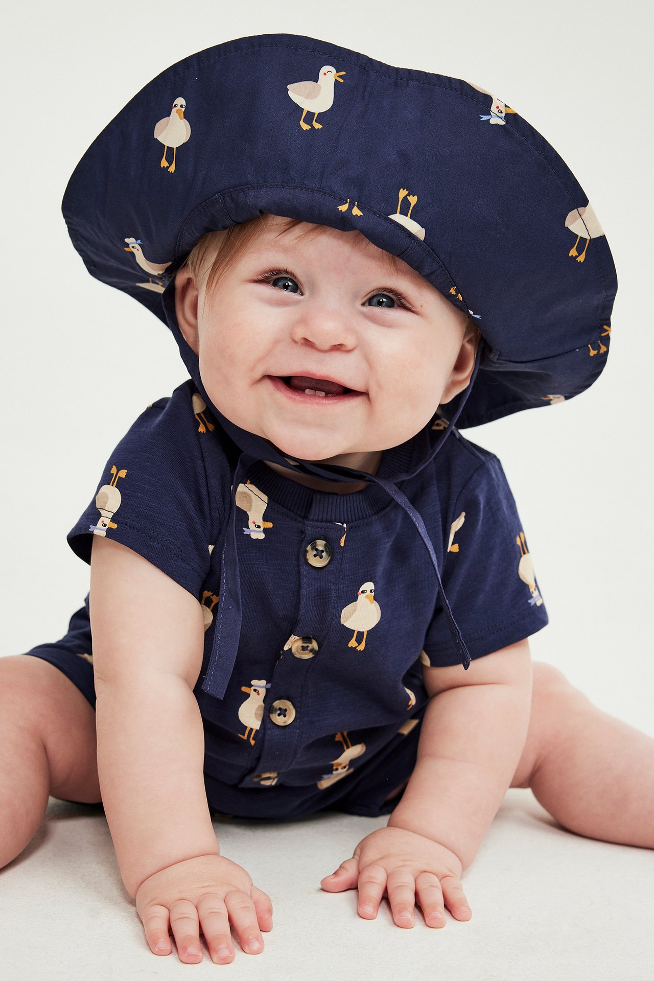 Hymyilevä vauva sinisessä ankka-kuvioisessa lyhythihaisessa romperissa napeilla ja yhteensopivassa hatussa.