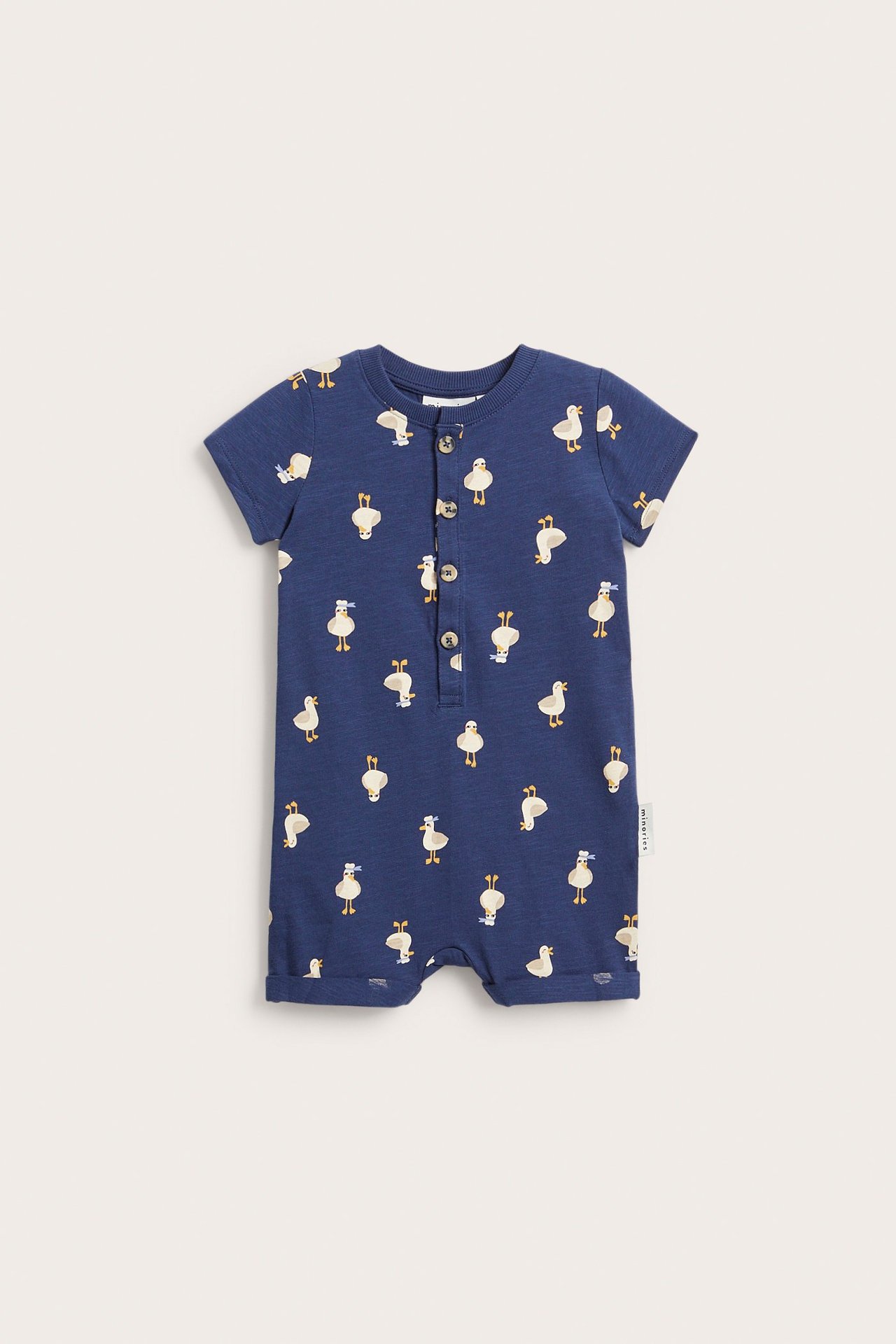 Widok z przodu niebieskiego, bawełnianego pajacyka niemowlęcego z krótkim rękawem, w kaczki, zapinanego na guziki.