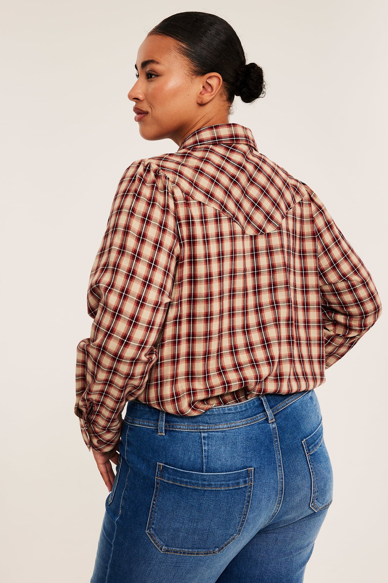 Kvinnlig modell i en beige och rödrutig långärmad skjorta med okdetaljer, stylad med blå jeans.