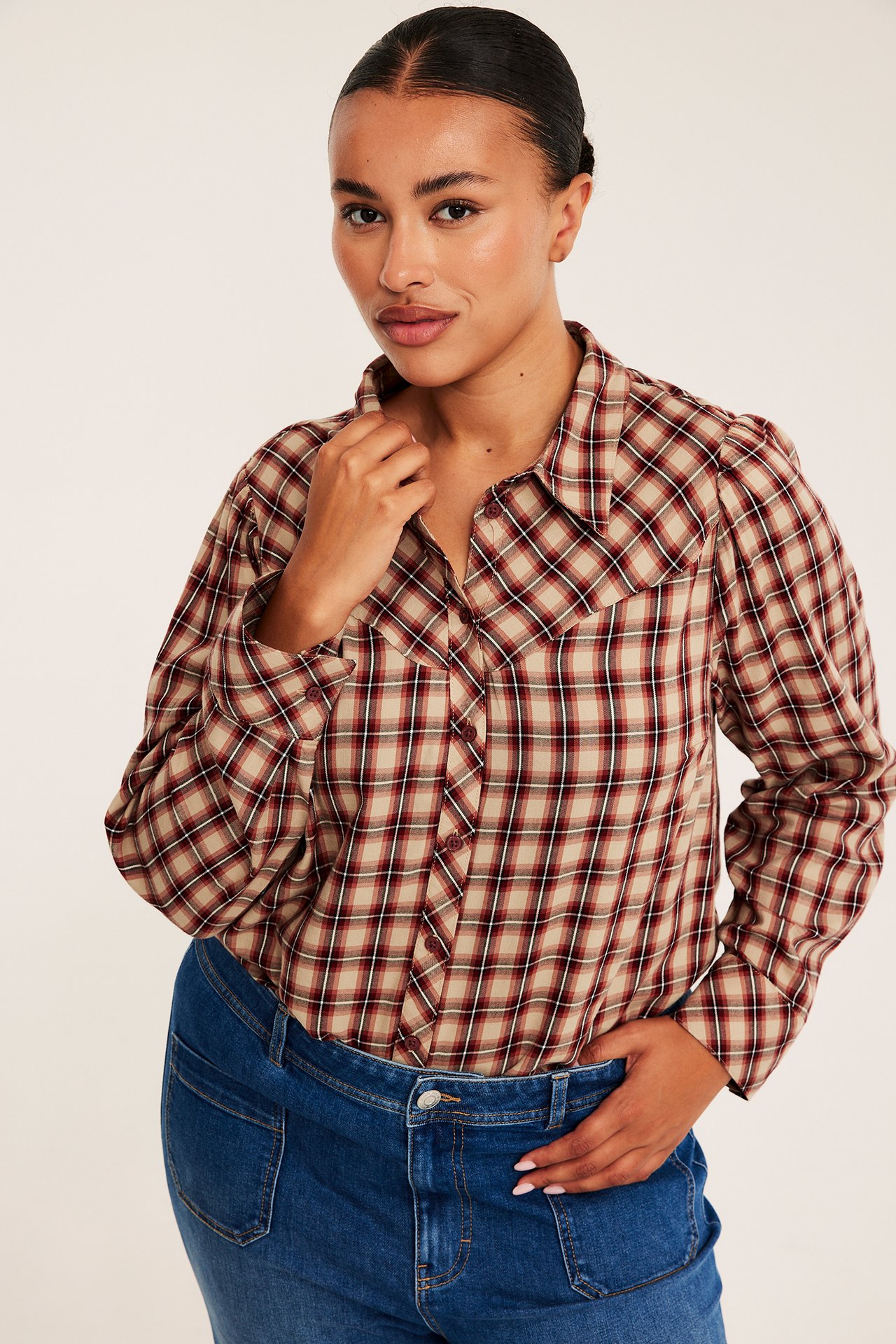 Frontvy av en kvinna i en rutig beige, röd och svart knappskjorta med blå jeans.